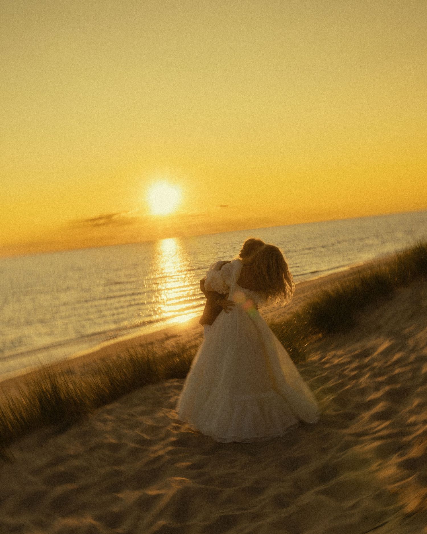 A bride in a white gown stands on sandy beach dunes at golden sunset with ocean in background.