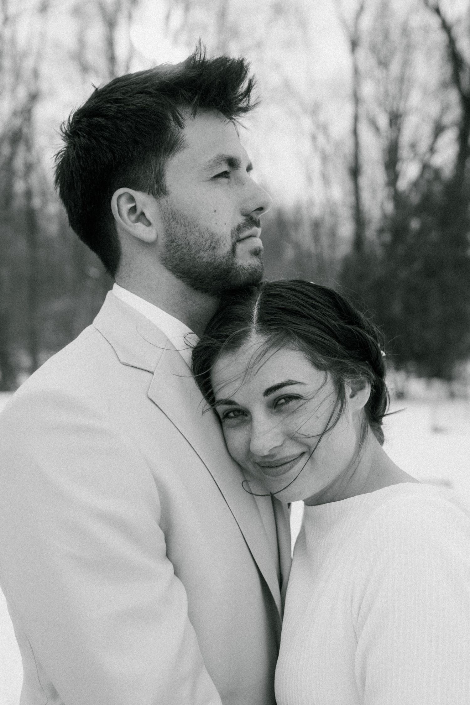 A romantic winter photoshoot with a couple in white outfits embracing against a snowy forest backdrop.