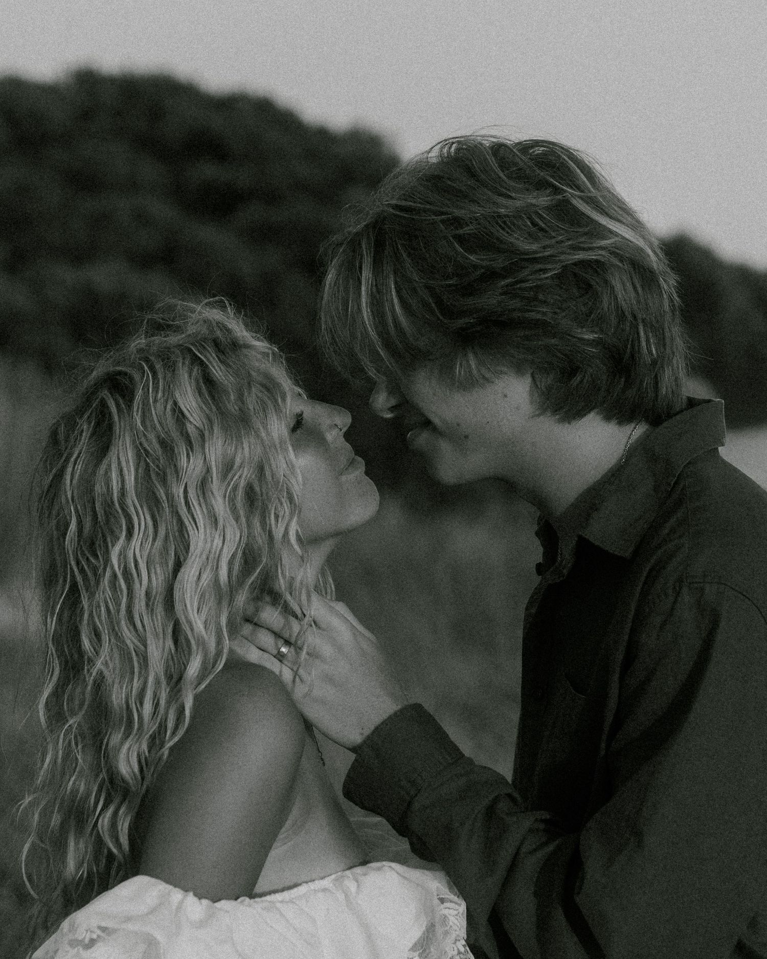 Black and white photo of a romantic couple almost kissing outdoors, woman with curly hair in white dress, man in dark shirt.