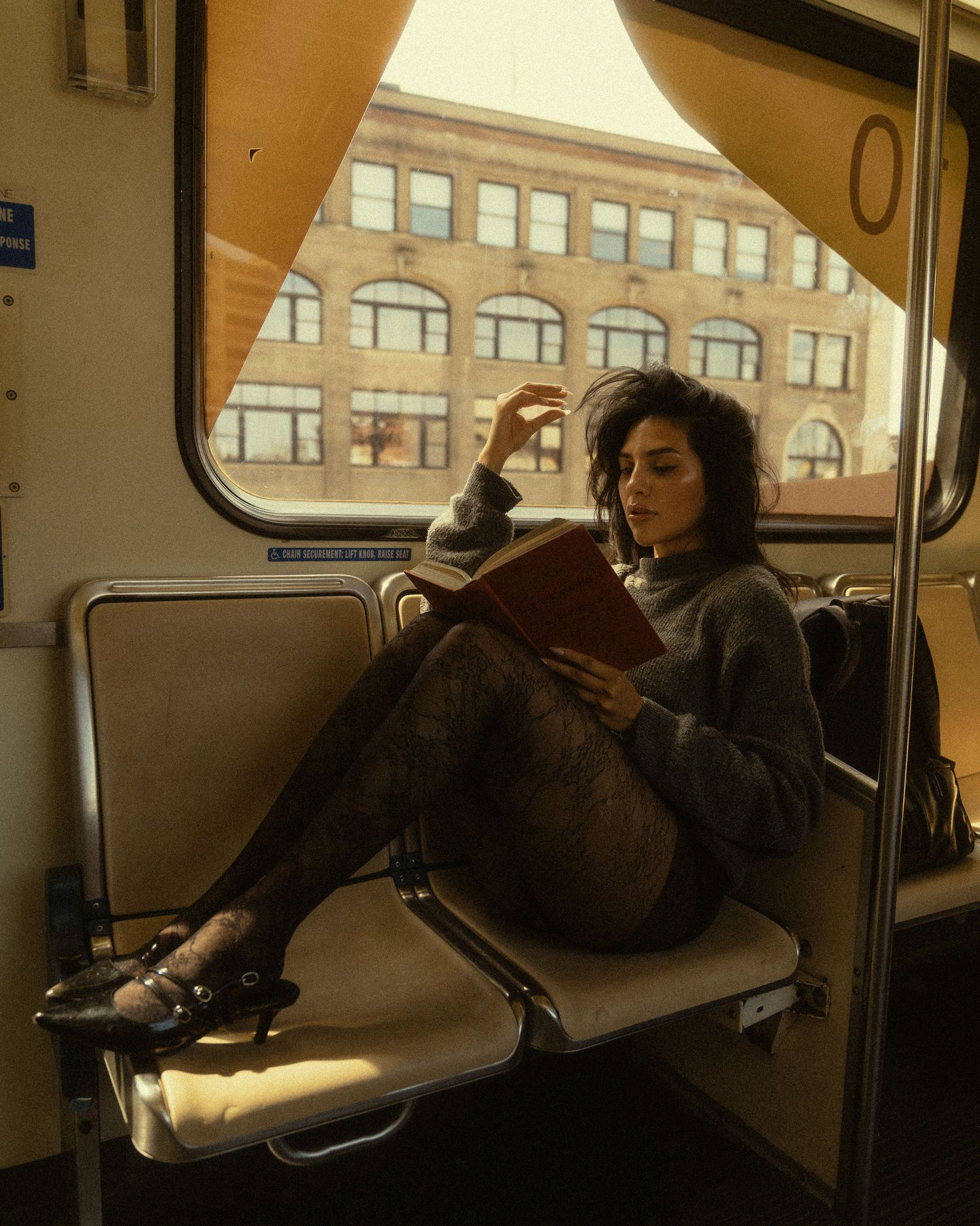 A young woman reads a book while sitting on a subway seat, wearing a cozy sweater and patterned tights.