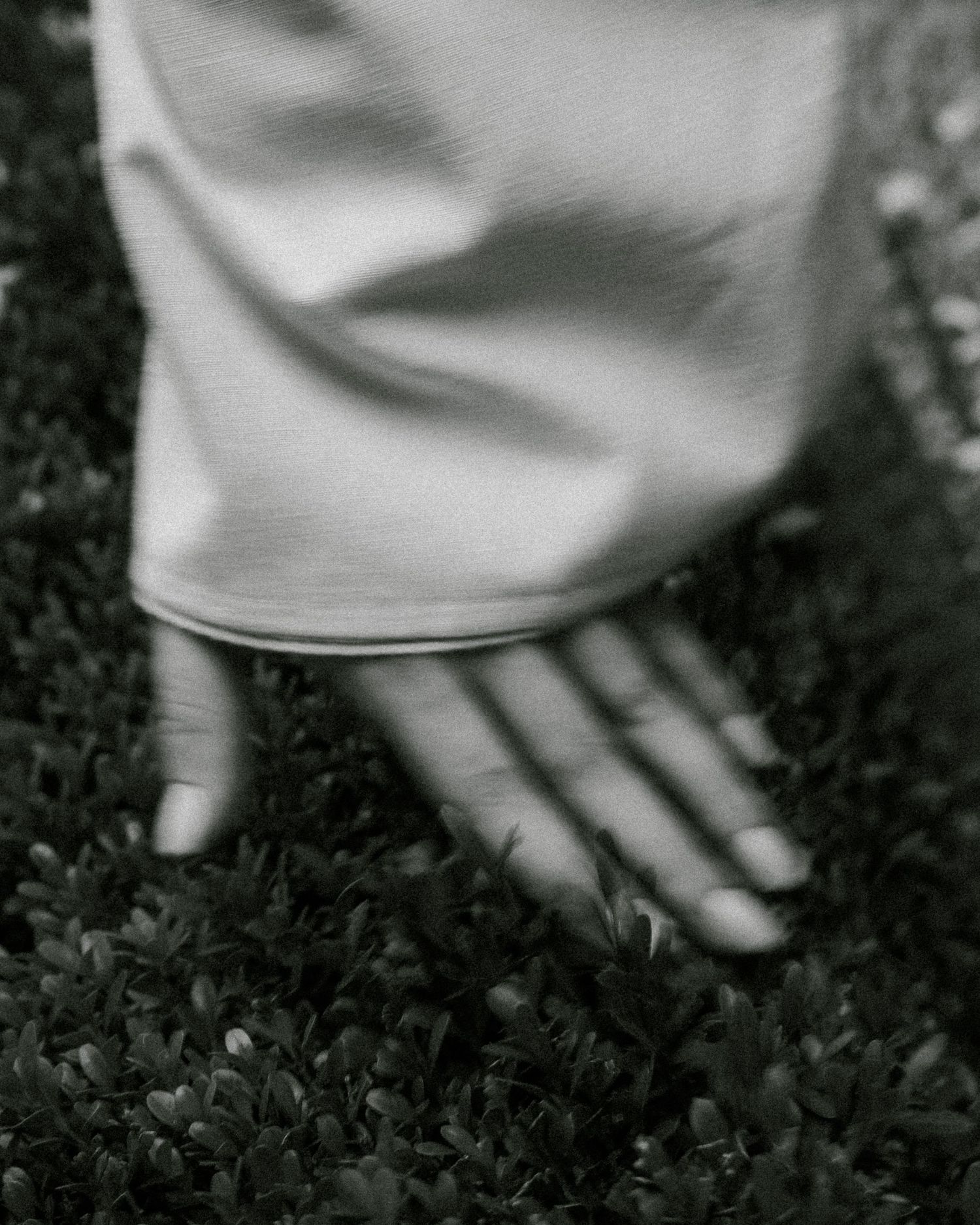 Black and white photo of a hand resting on lush green grass beside a flowing white dress hem.