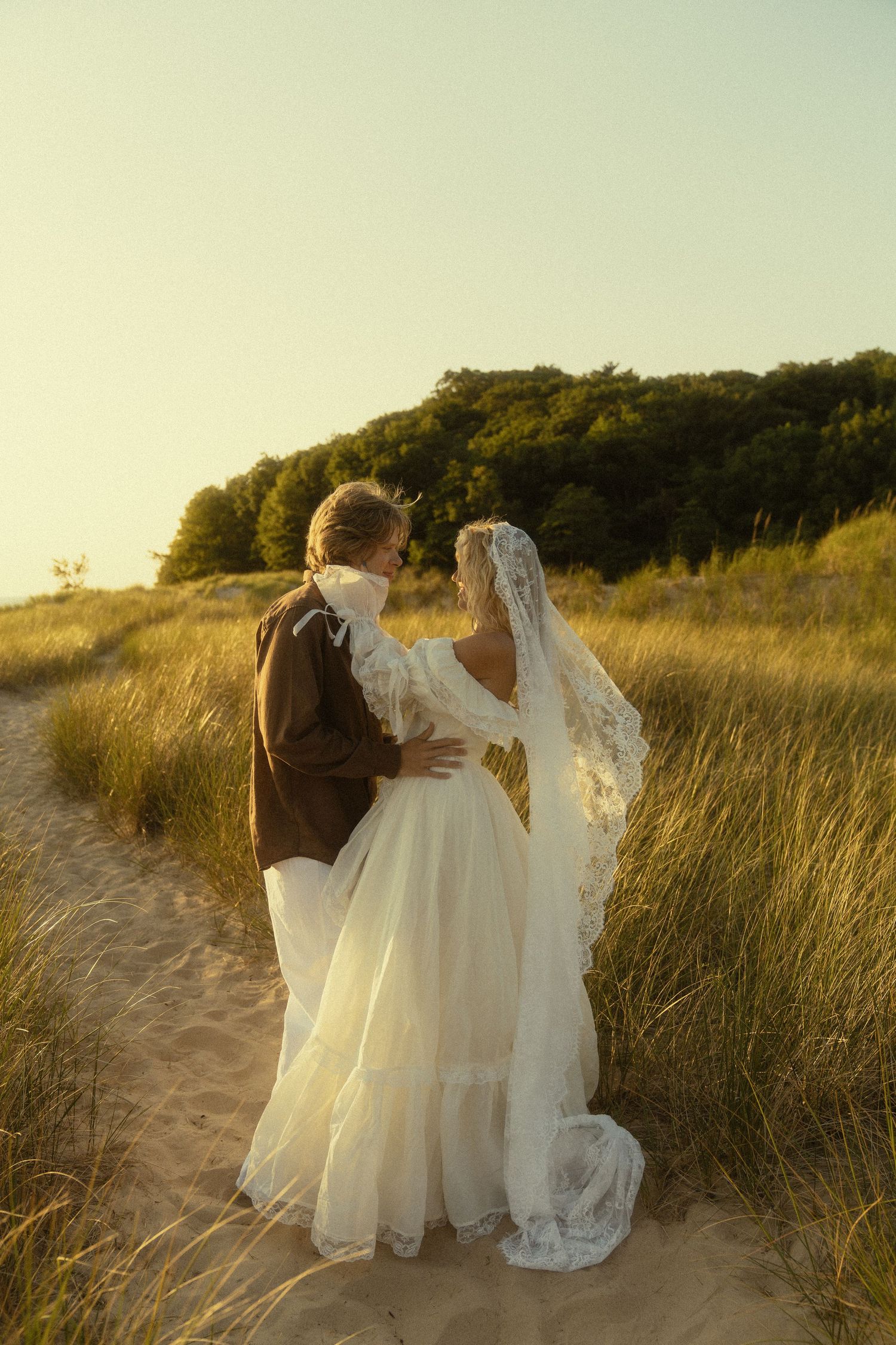 A bride and groom embrace on a sandy path surrounded by golden grass and trees during a romantic sunset wedding.