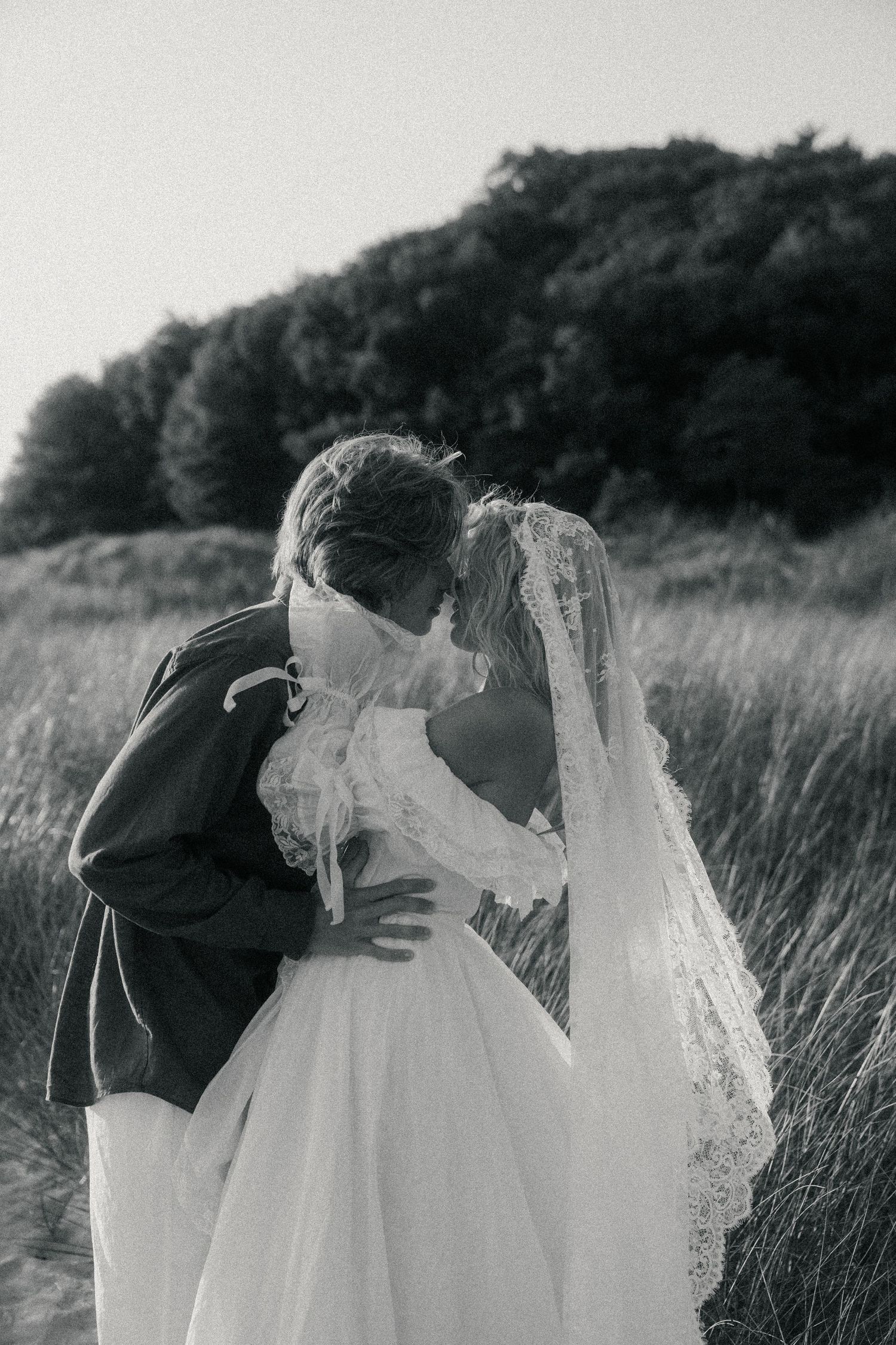 Black and white photo of a bride and groom sharing a kiss outdoors, with tall grass and trees in the background.