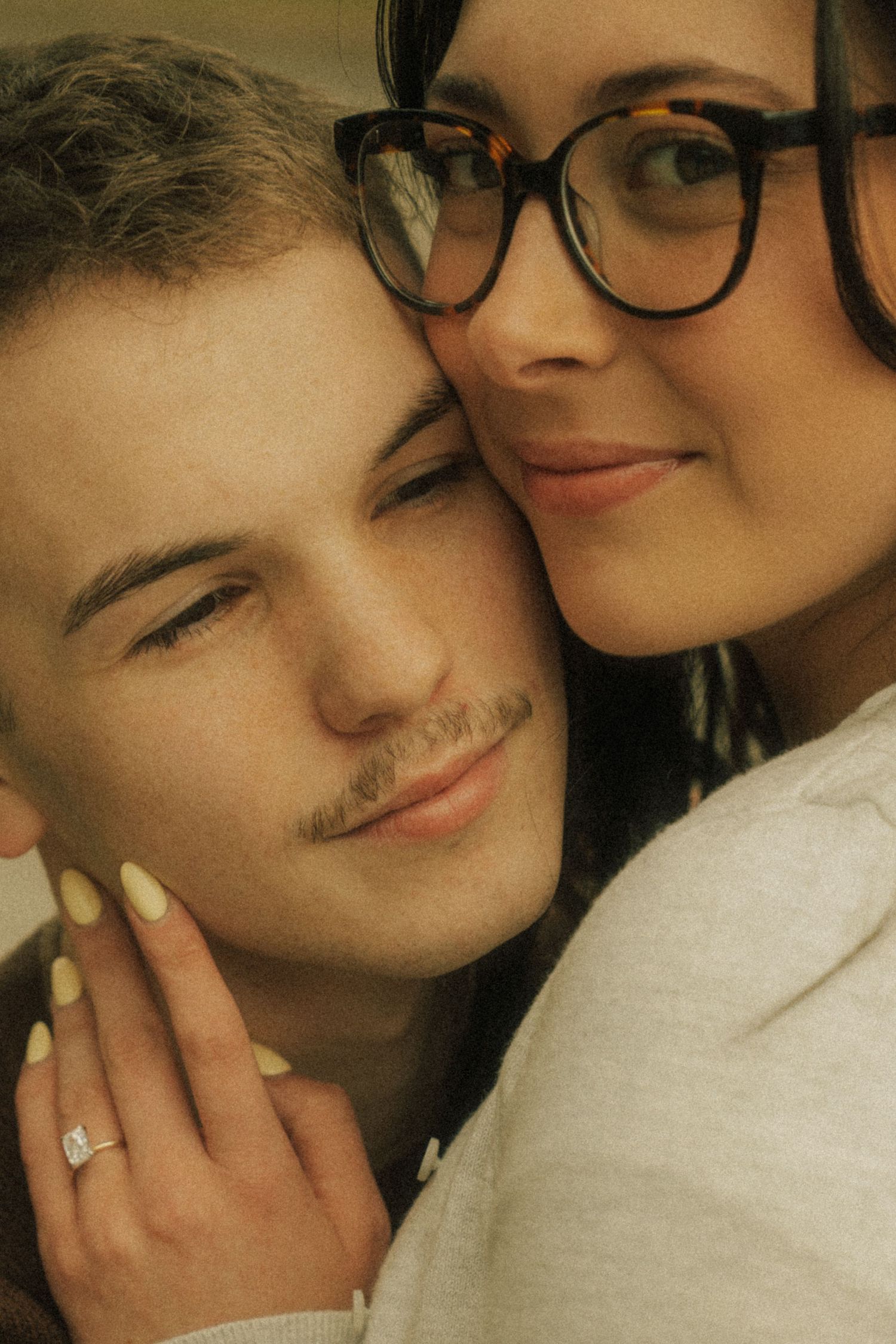 Couple posing closely together, woman wearing glasses and engagement ring resting her cheek on man's head.