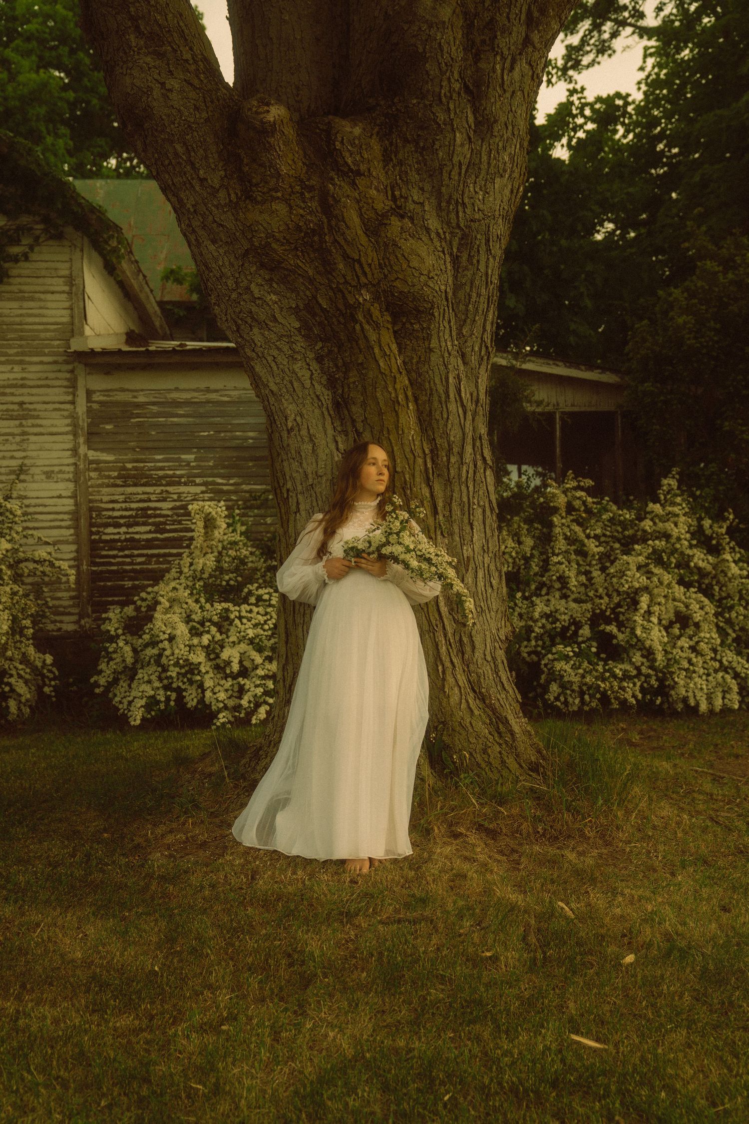 Woman in white dress holding flowers stands barefoot by large tree near rustic building at dusk.
