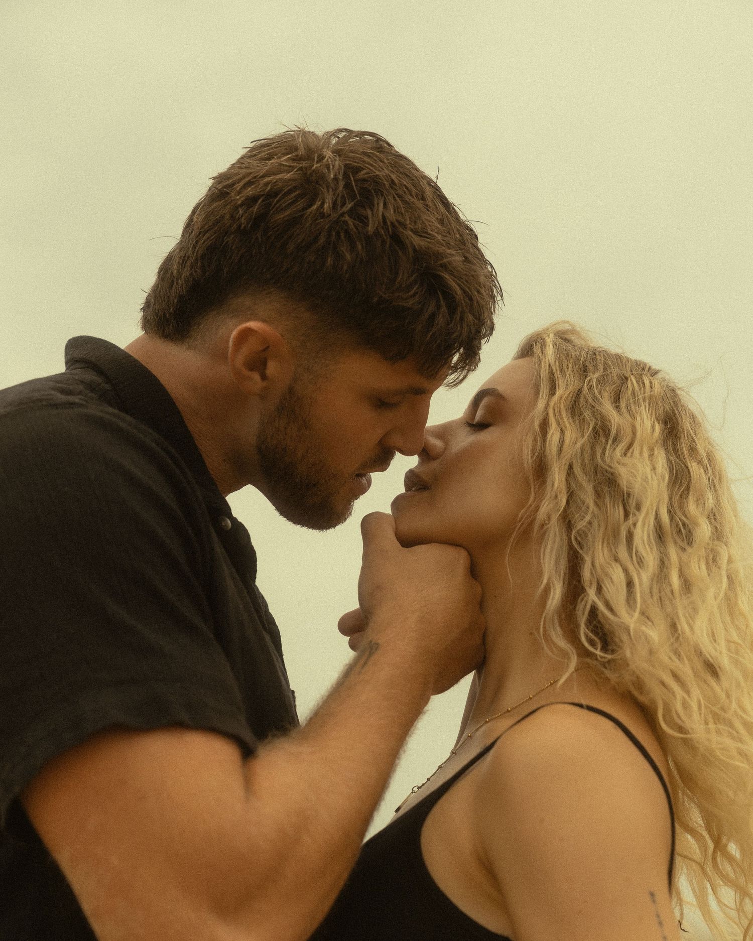 A man and woman with curly blonde hair share a romantic almost-kiss moment against a soft, misty background.