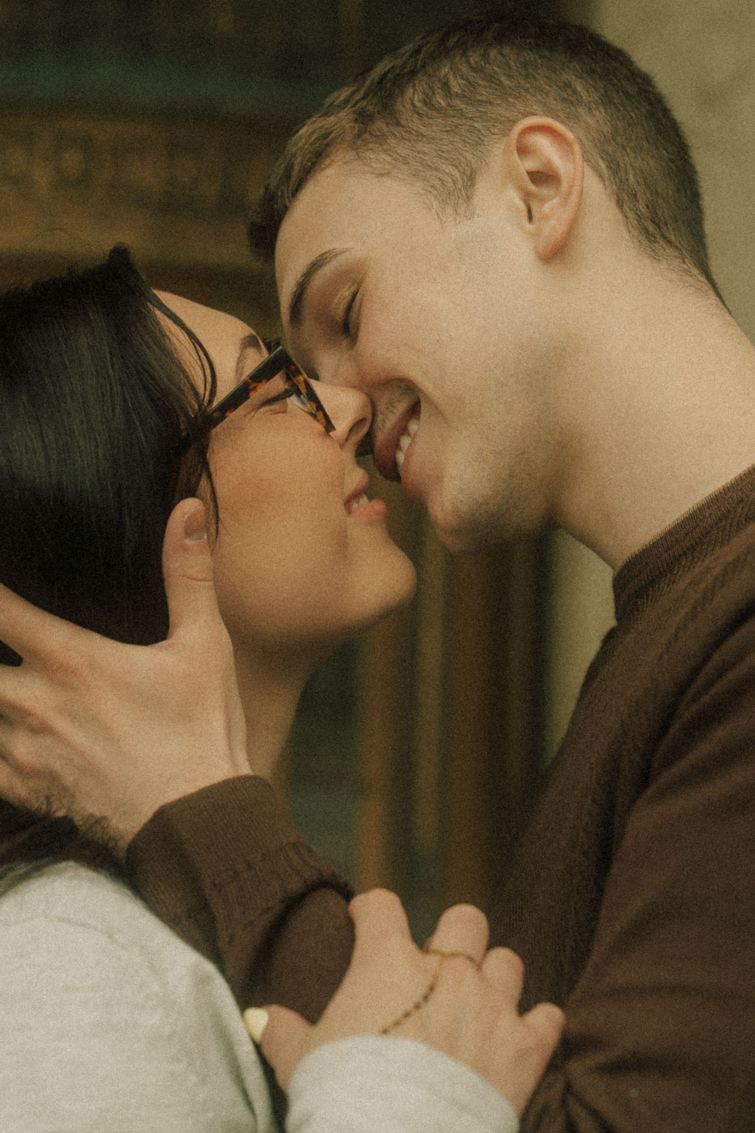 A couple sharing a romantic moment, smiling and about to kiss, the woman wearing glasses and the man in a brown sweater.