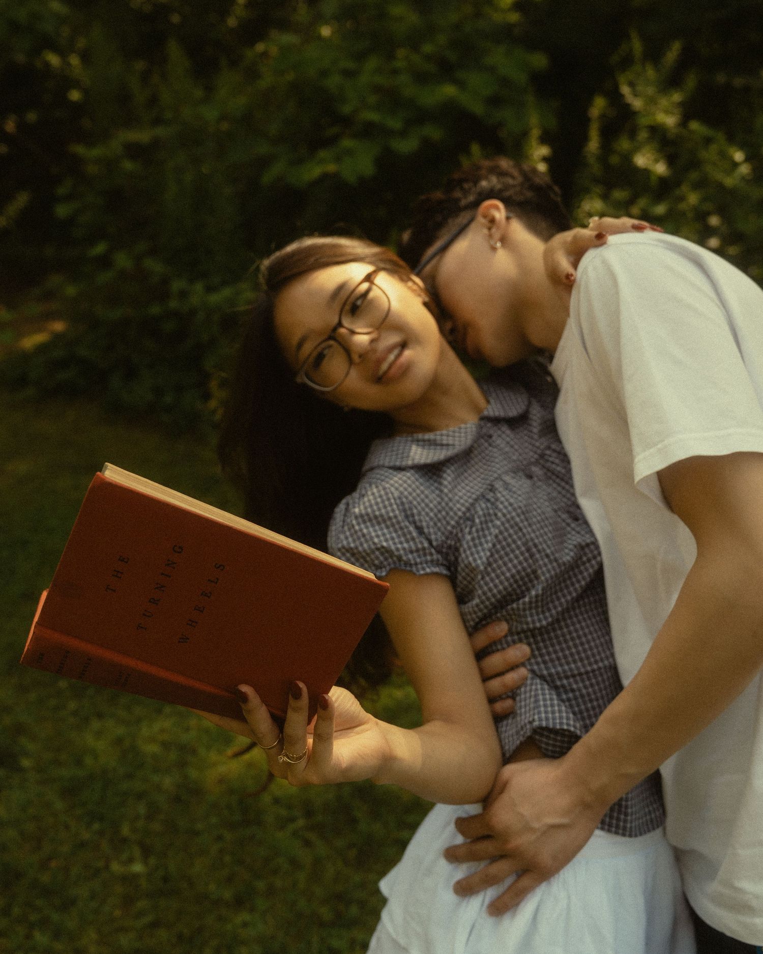 Couple embracing outdoors, woman in glasses holding a red book while man kisses her cheek in a lush garden.