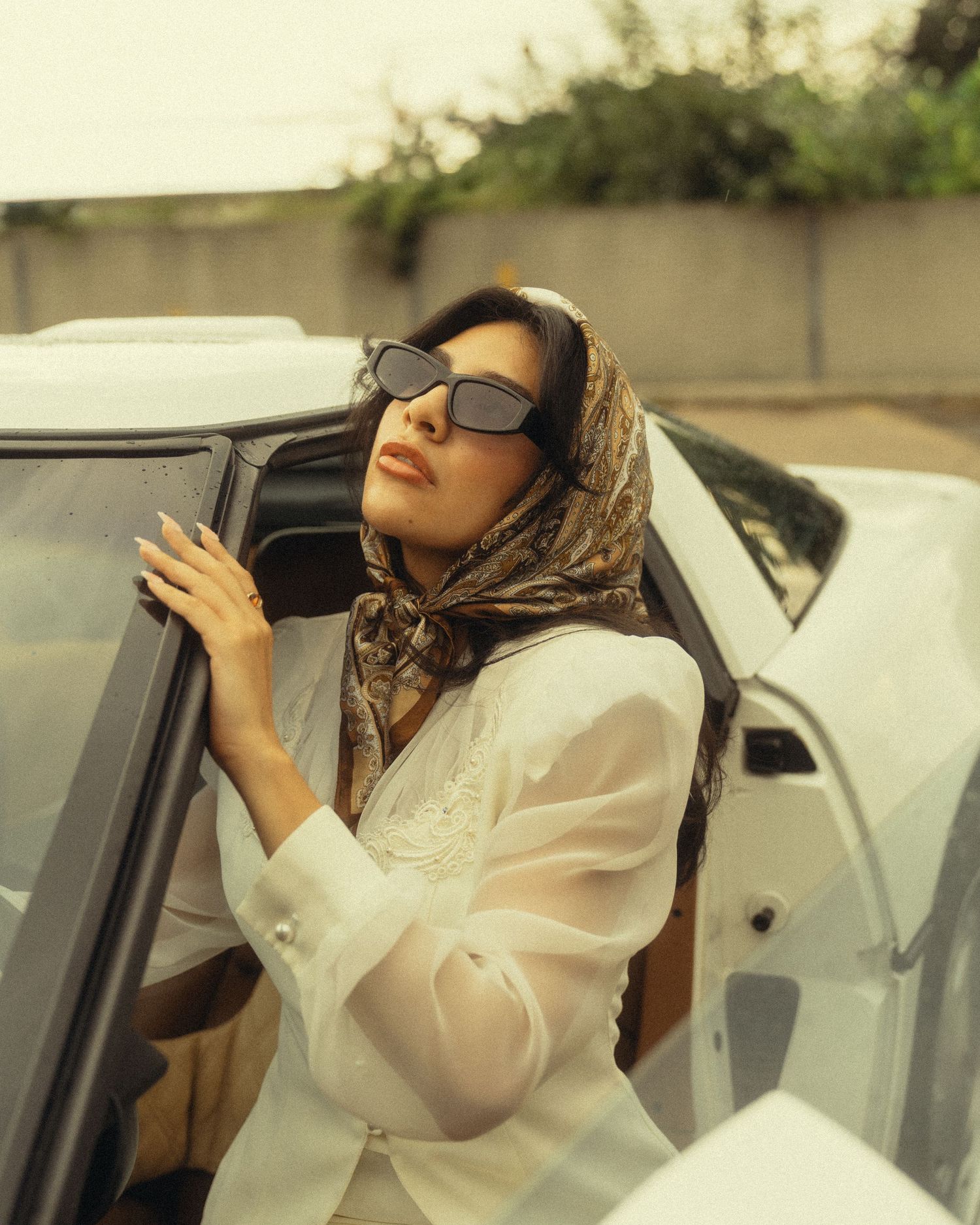 Woman in white blouse, patterned headscarf, and dark sunglasses leaning on a white car door outdoors.