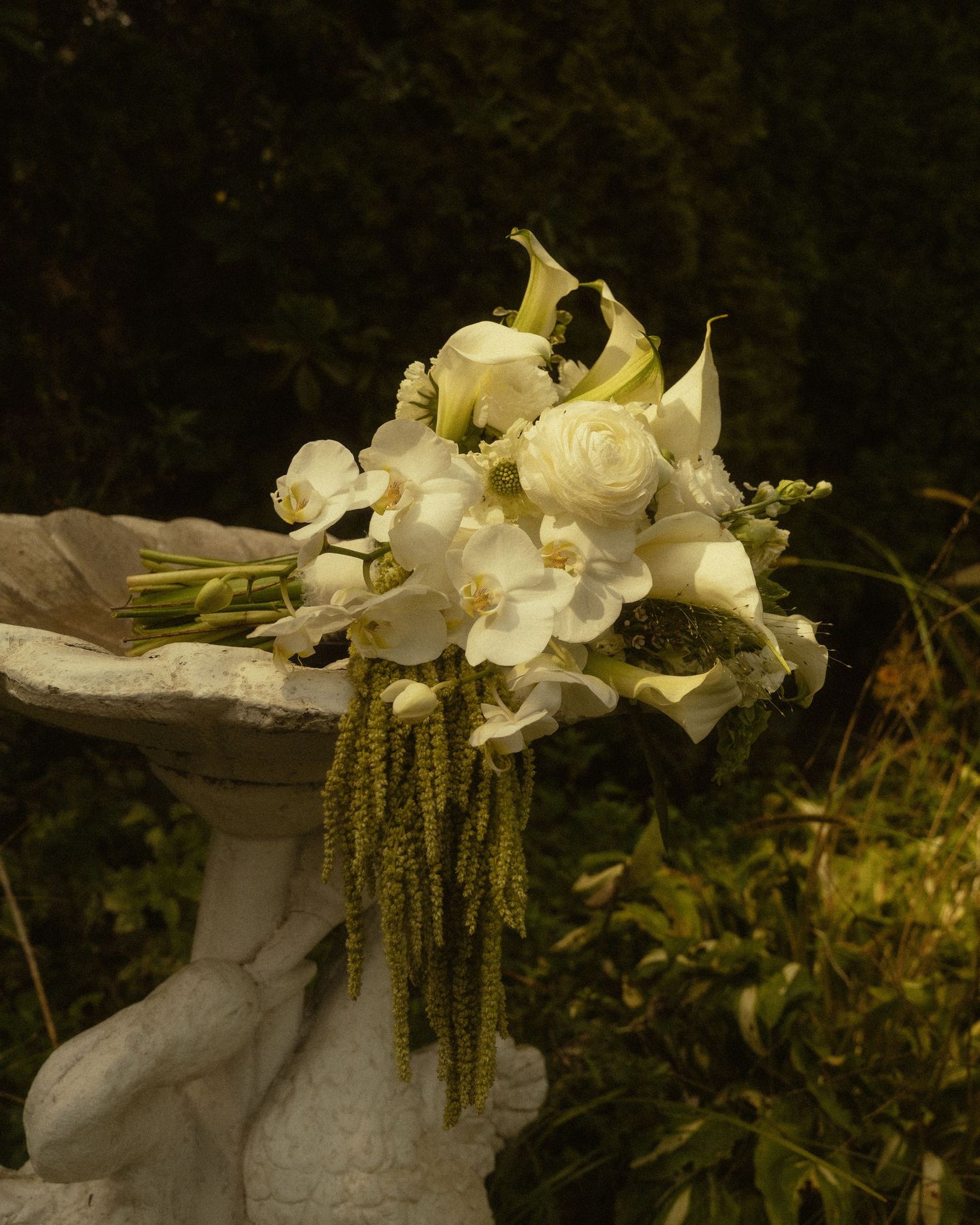 Elegant white wedding bouquet with calla lilies, roses, and orchids resting on a stone birdbath in a garden.