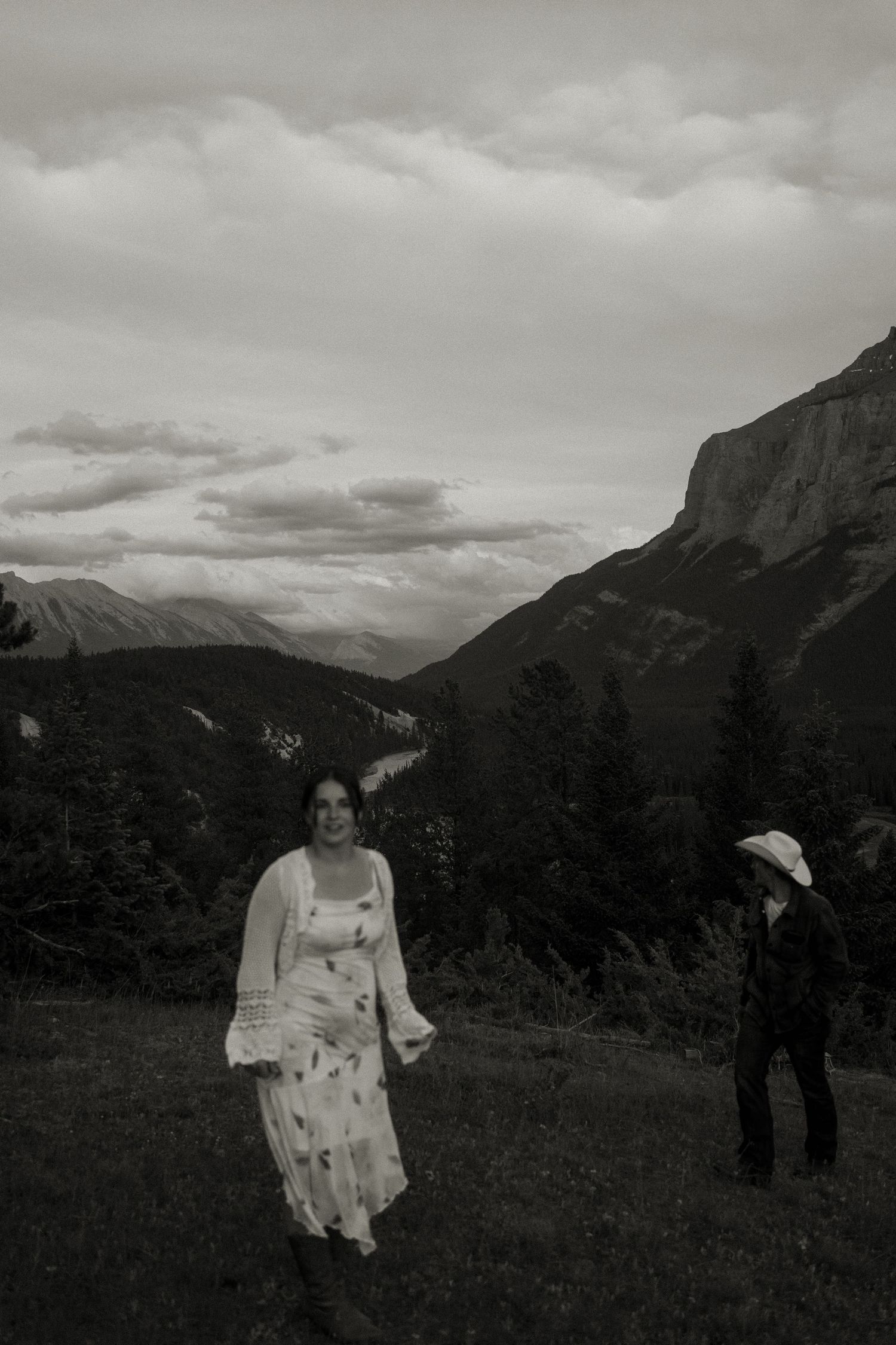 A black and white portrait of a couple in a mountain meadow with dramatic peaks behind them.