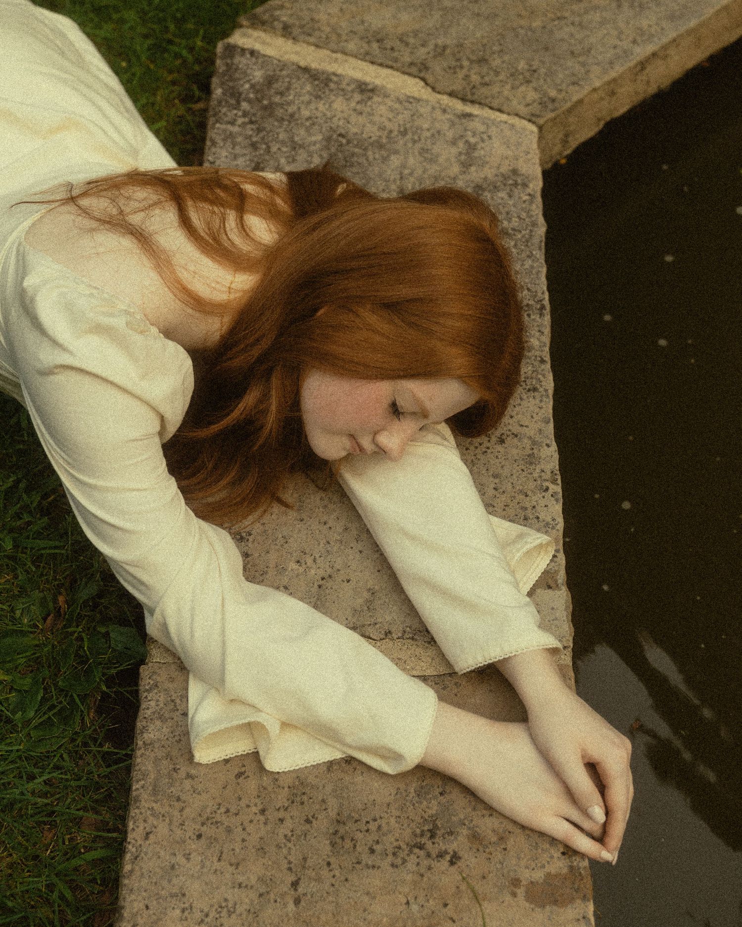 A red-haired woman in a white dress lies on a stone ledge, reaching her hand toward a dark reflective pool.