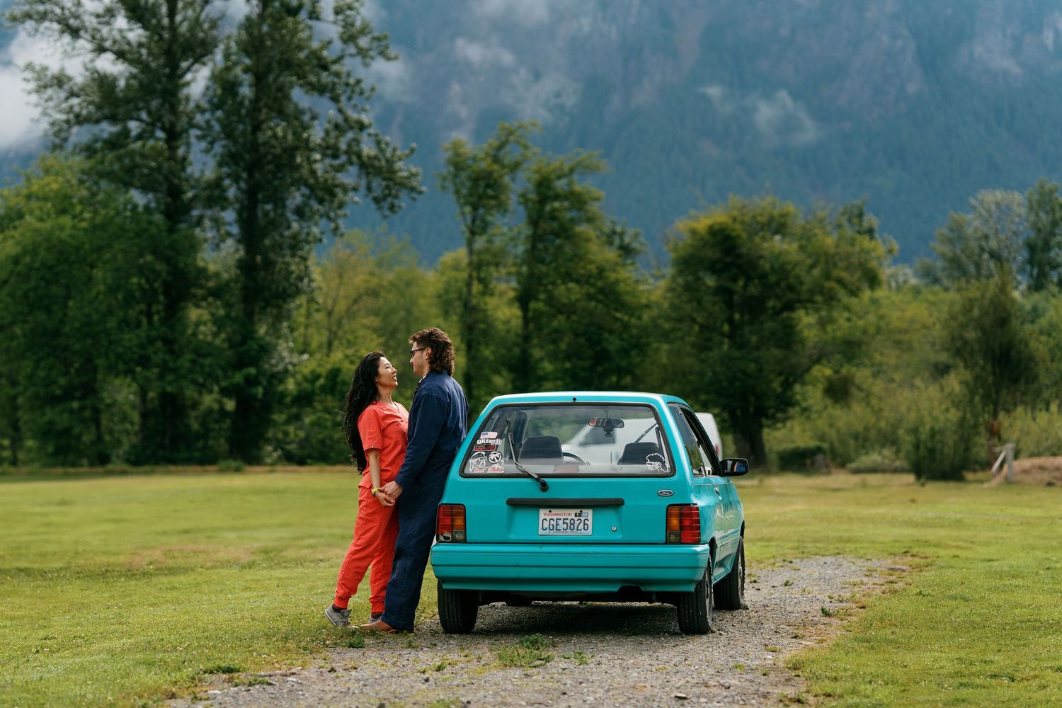 Couple standing together by a teal vintage car in a scenic green field with mountains in the background.
