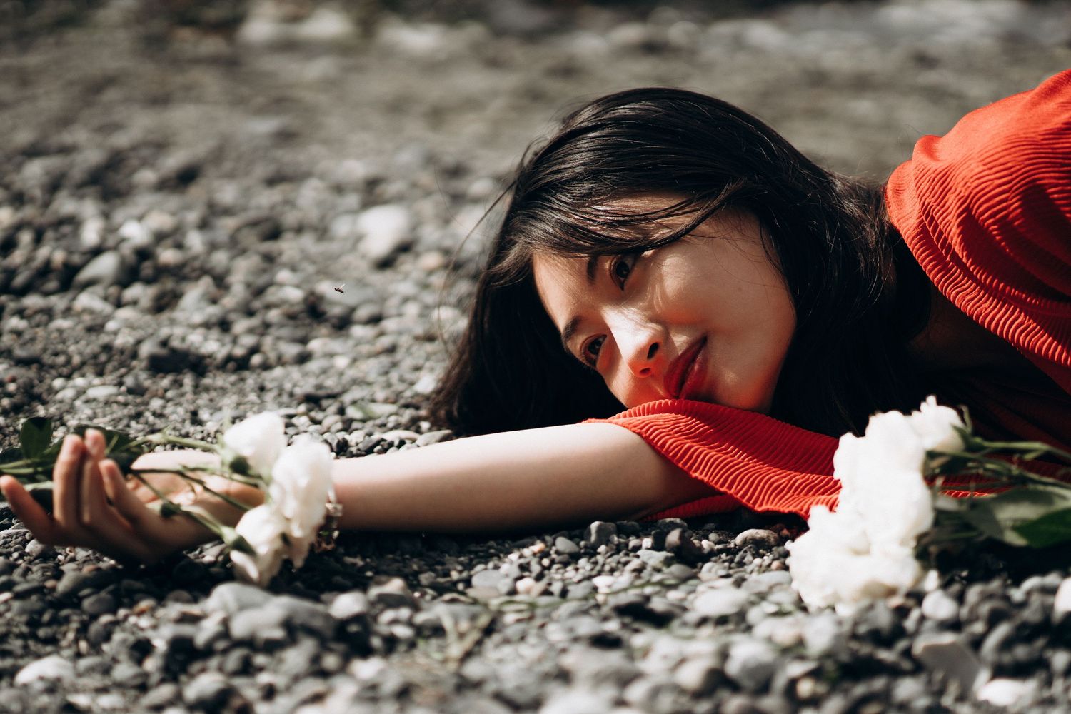 Young woman in red dress lying on pebbles reaching toward white roses.