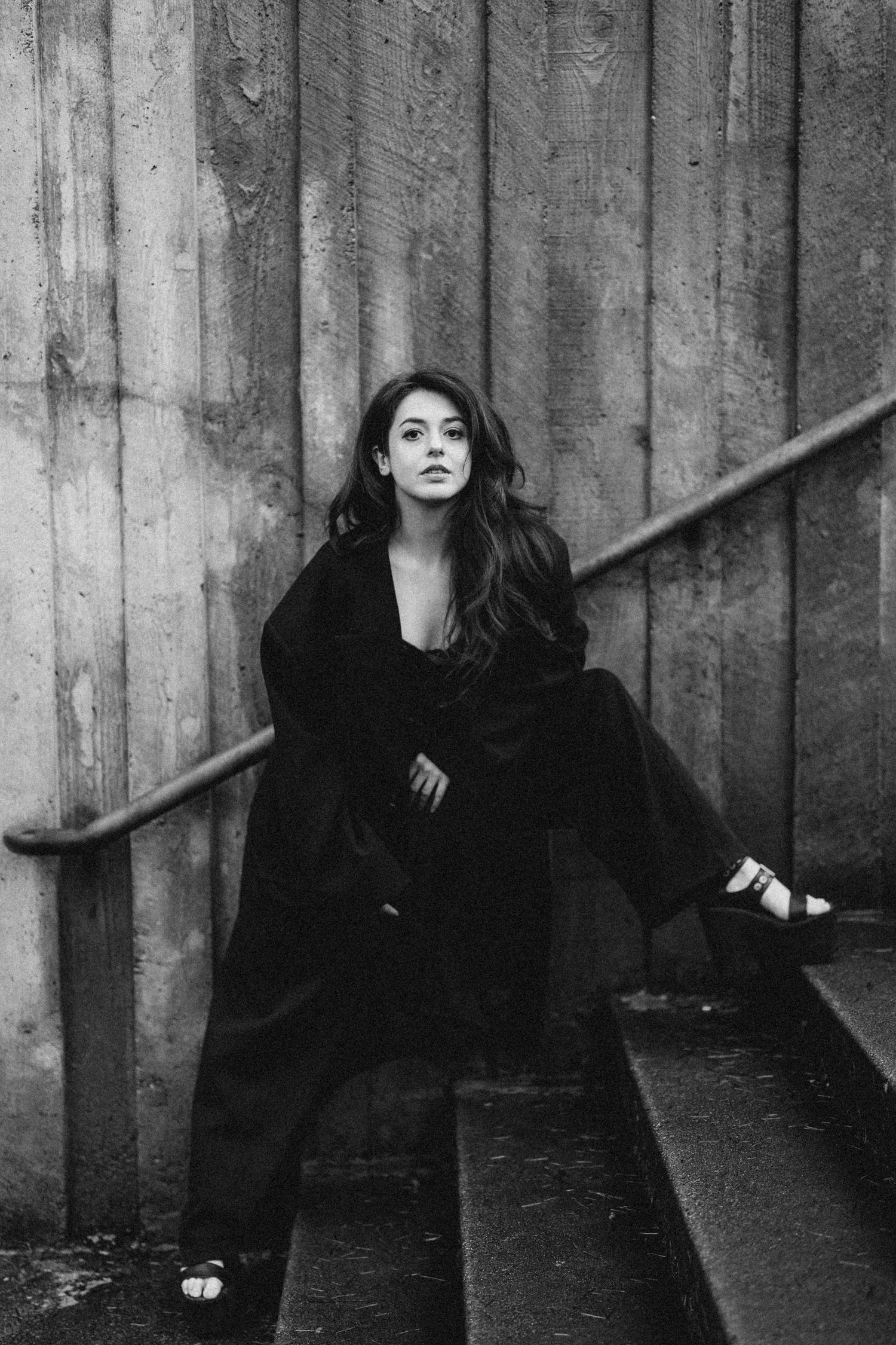 Black and white photo of woman in long black coat sitting on stairs against wooden wall.