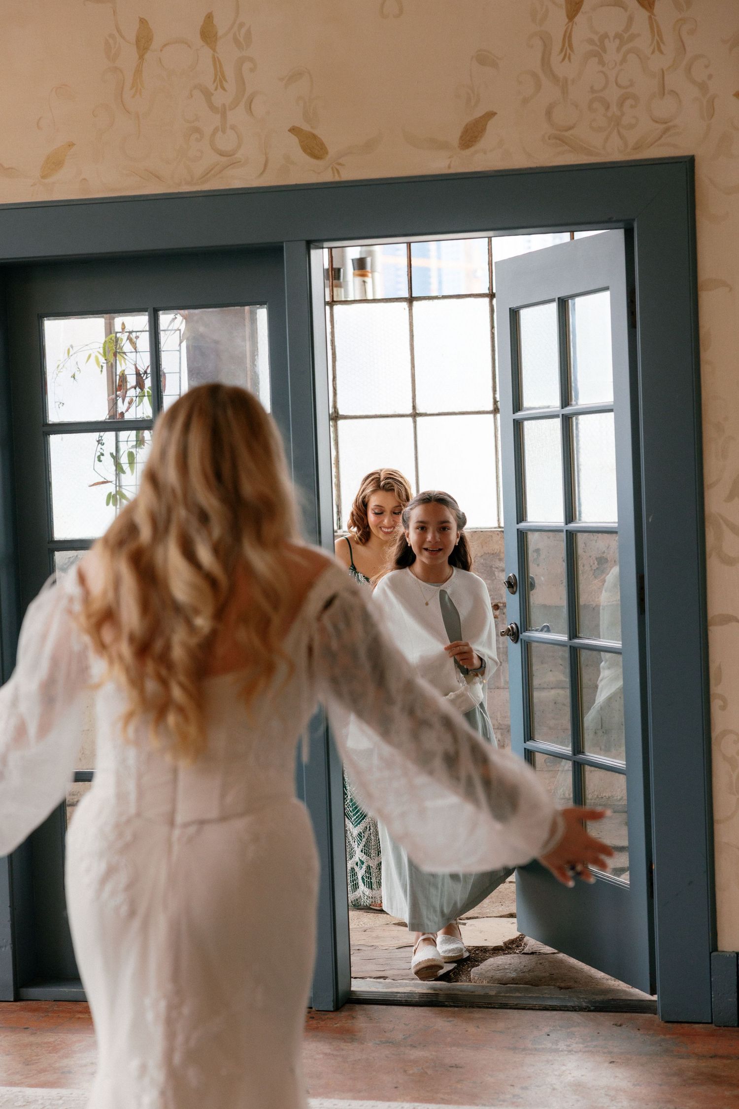 Bride opens arms to welcome flower girl and bridesmaid through blue French doors.