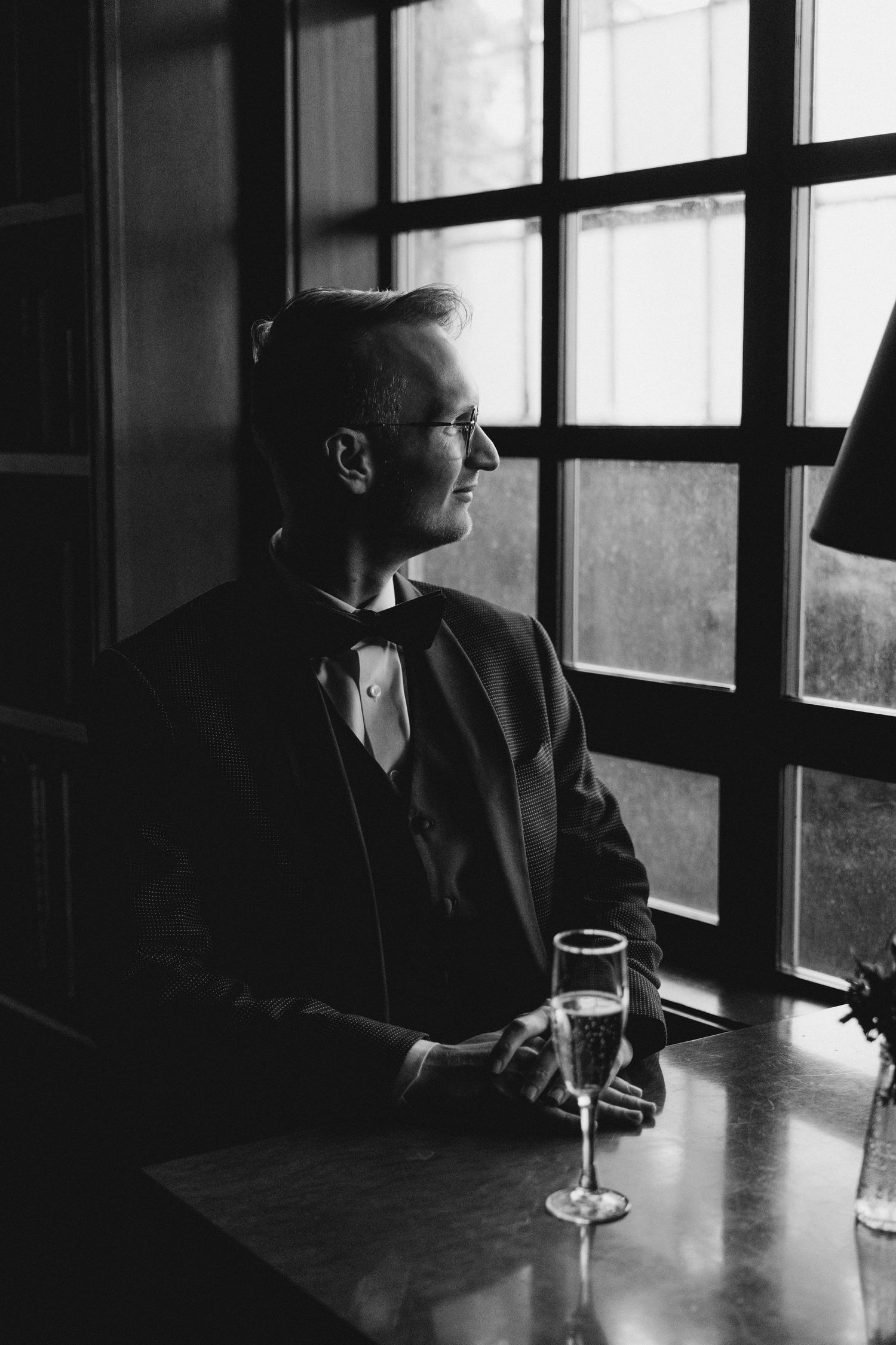 Black and white portrait of dapper groom in tuxedo with bow tie gazing out window with champagne.