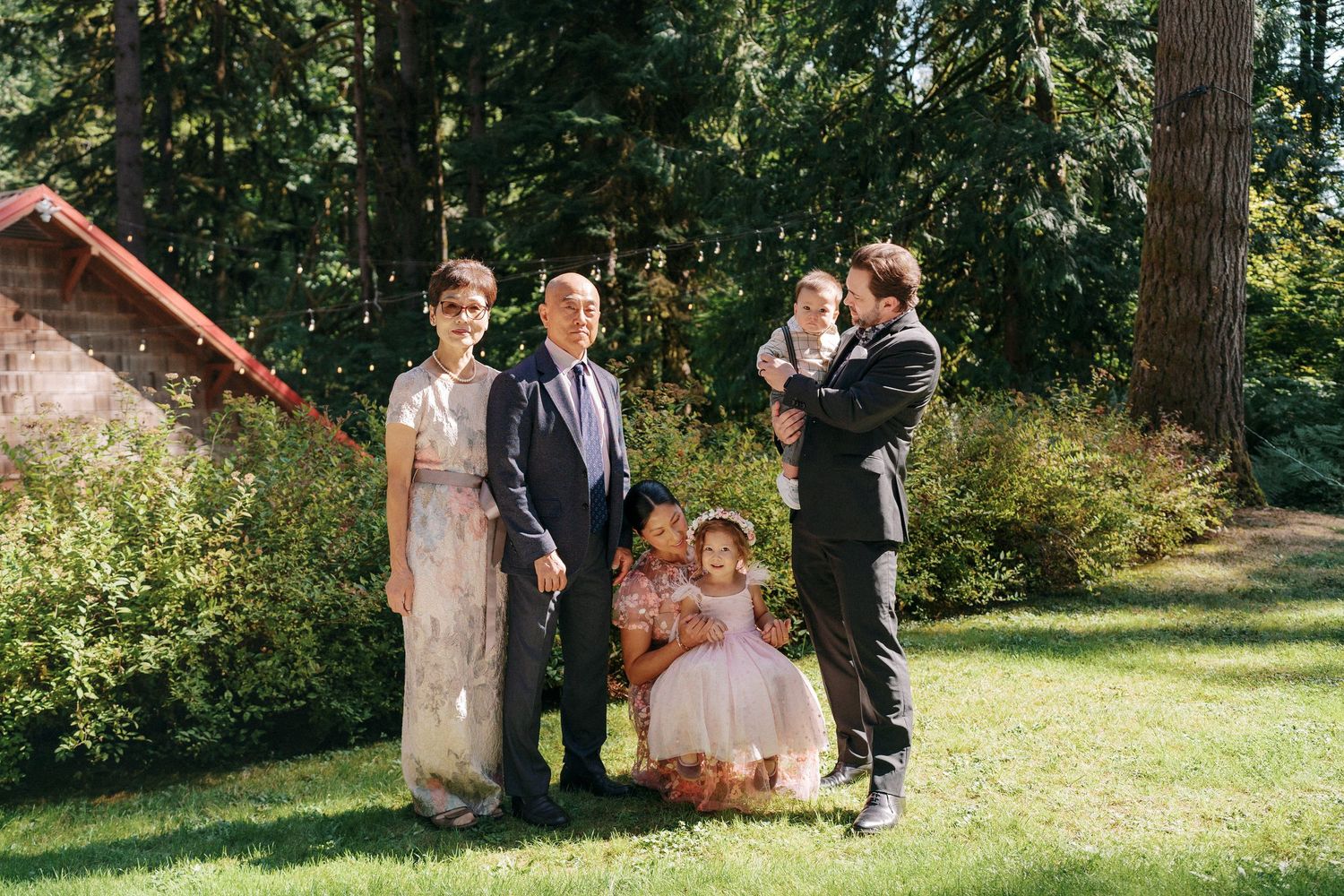 Family group portrait with children on a sunny lawn with tall evergreen trees behind.