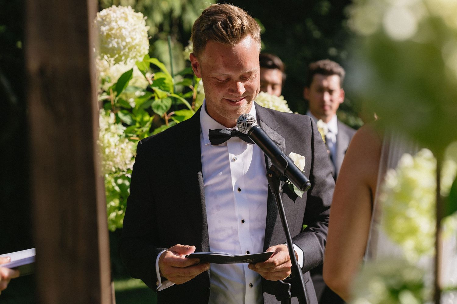 Emotional groom reading wedding vows at outdoor ceremony wearing black tuxedo with bow tie.