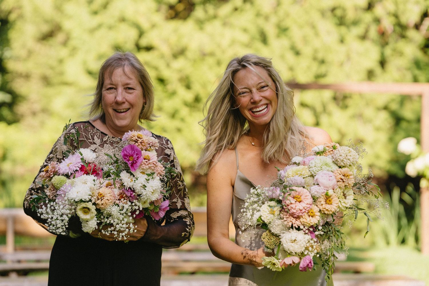 Two women smiling and holding colorful bridal bouquets with roses and wildflowers outdoors.