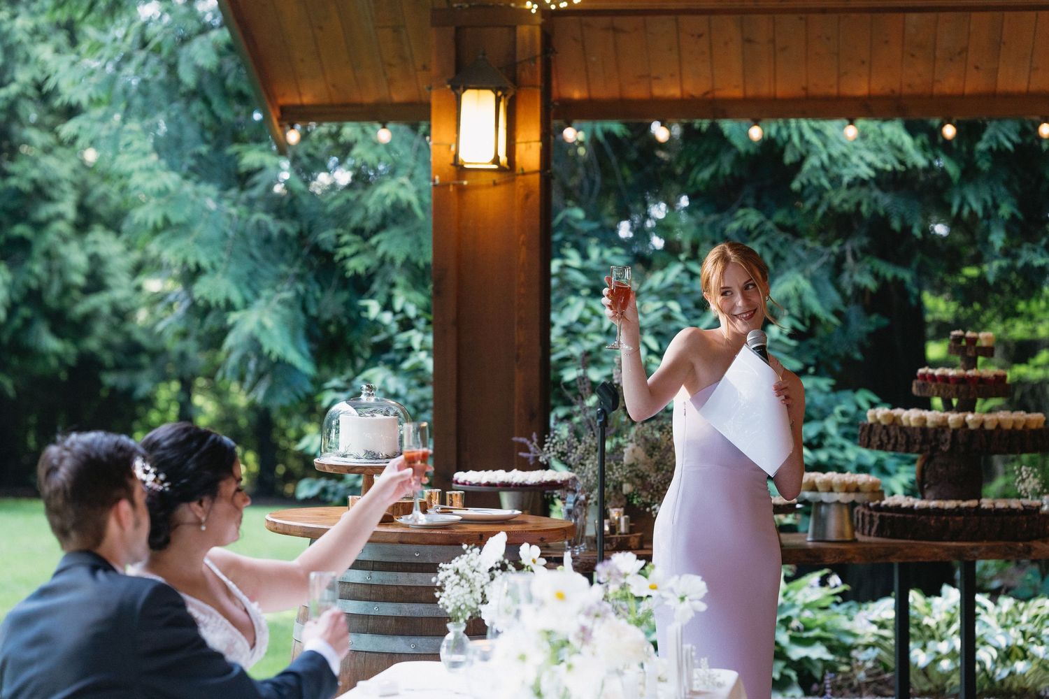 Bridesmaid in lavender gown raising a toast at elegant outdoor wedding reception under string lights.