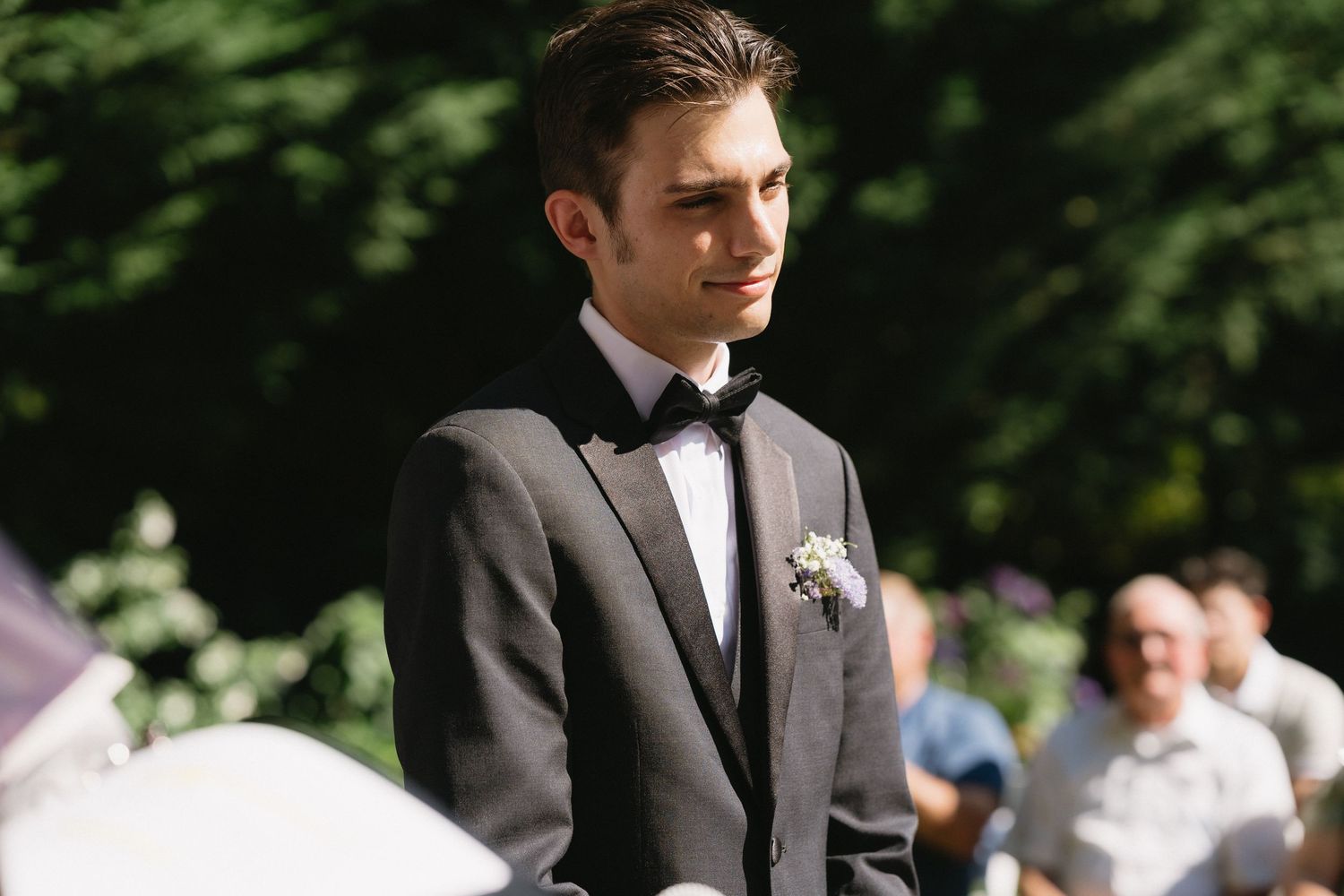 Handsome groom in gray suit with black bow tie and boutonniere stands at outdoor wedding altar.