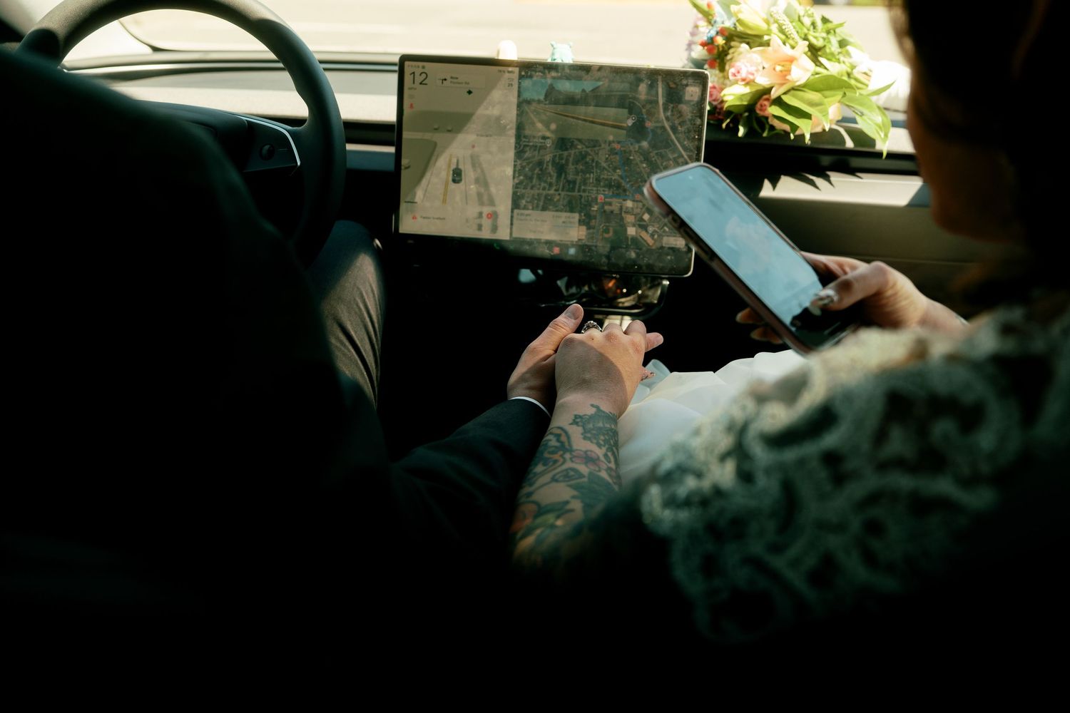 Bride and groom with tattoos using phone and Tesla touchscreen navigation in car.