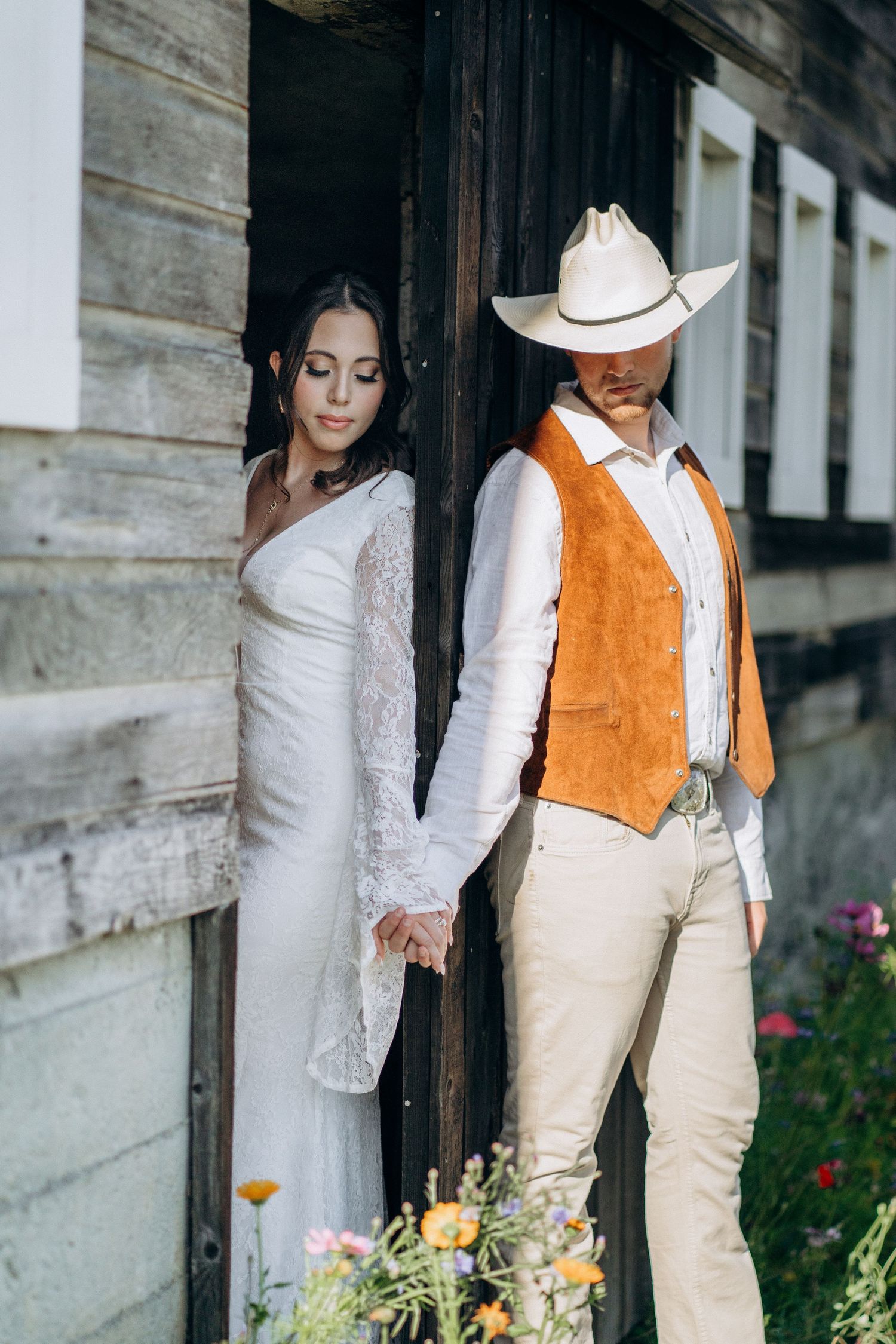 Bride and groom holding hands in barn doorway, groom wearing cowboy hat and suede vest.