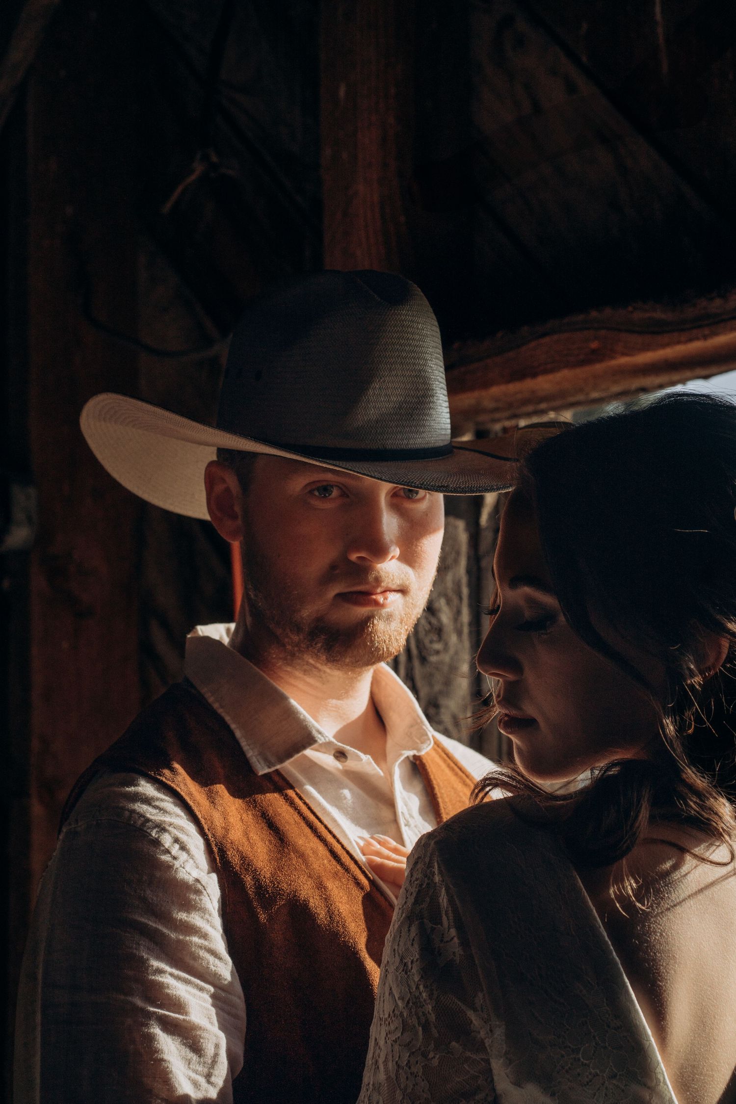 Groom in white cowboy hat and brown vest stands close to bride in dramatic warm barn light.