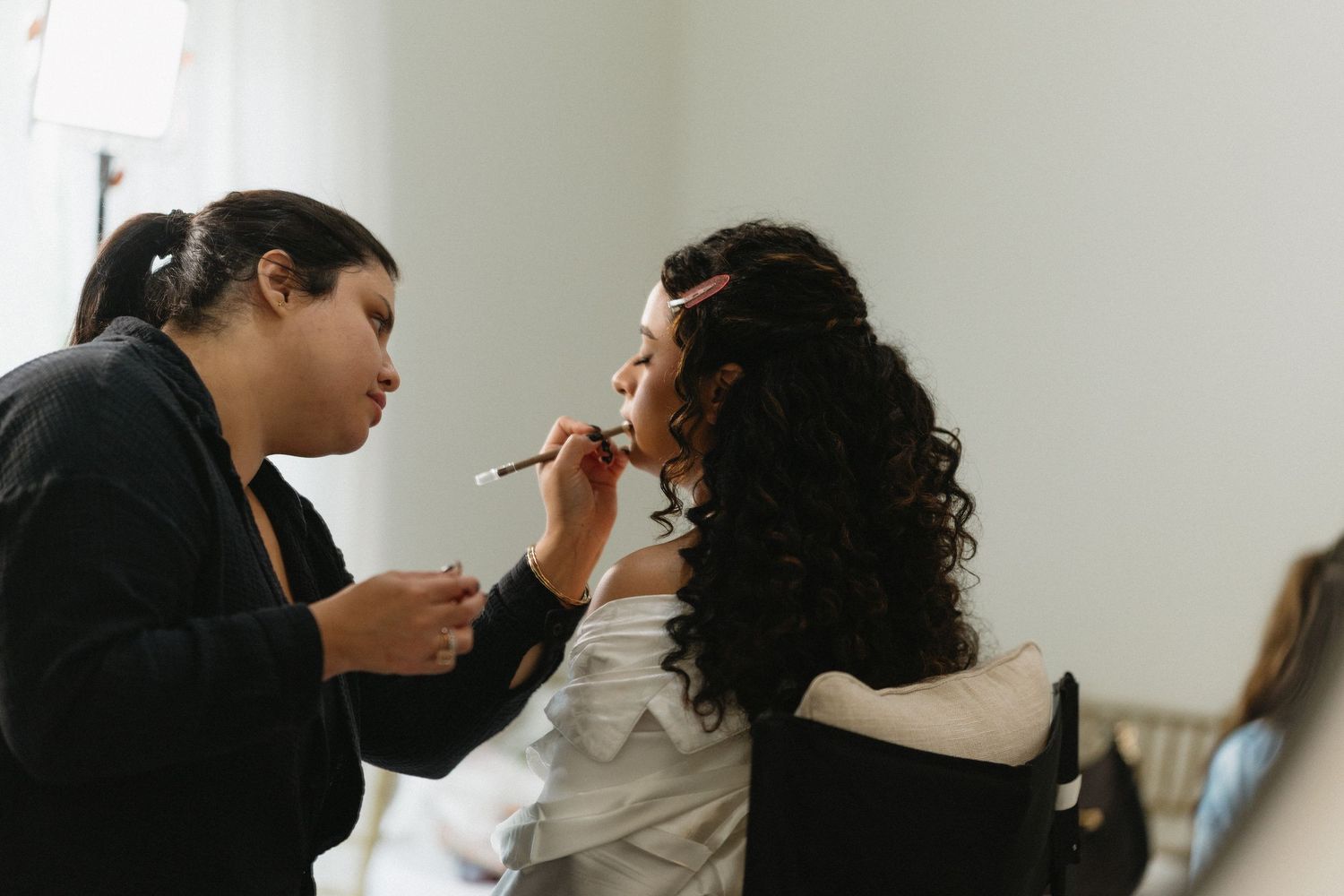 Professional makeup artist applying lipstick to curly-haired bride during wedding preparation.