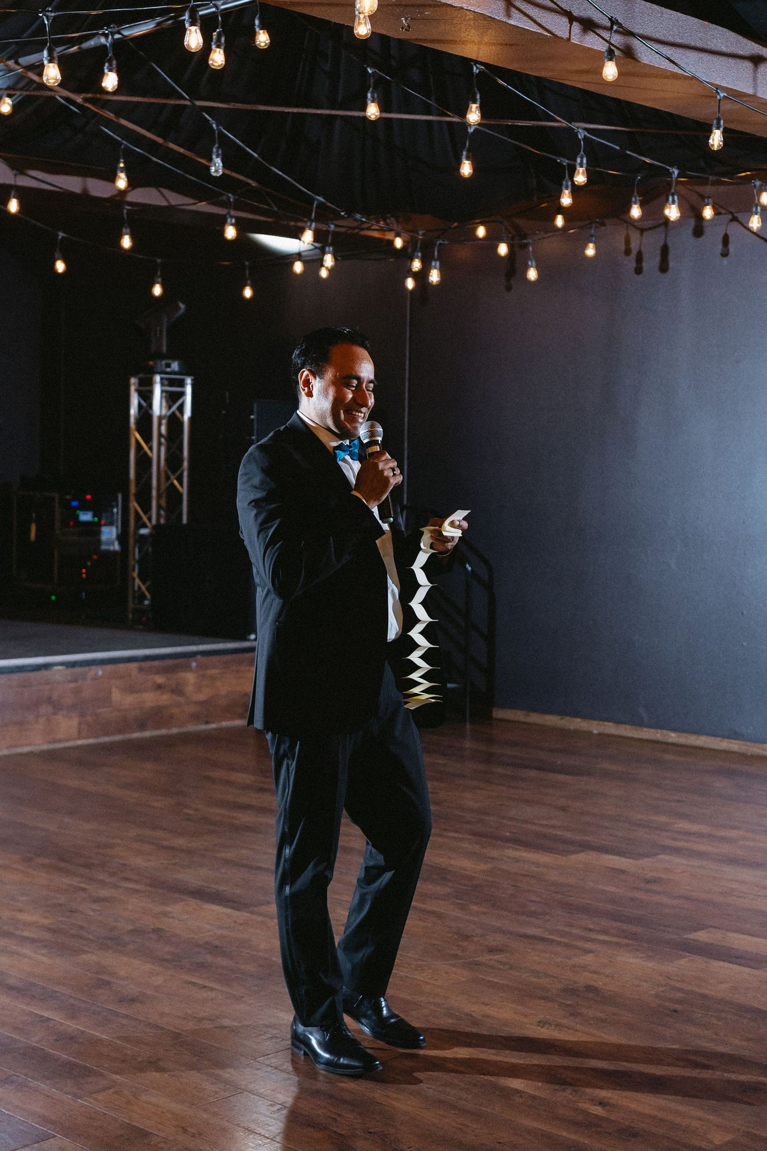 Man in black tuxedo giving speech with microphone on dance floor at wedding.