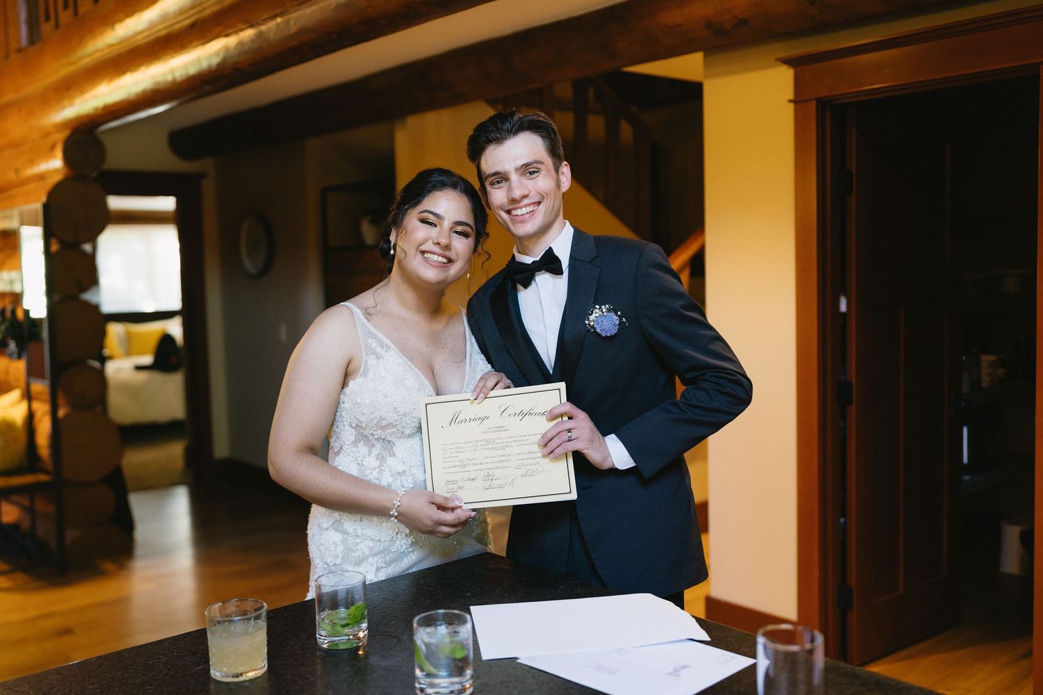 Smiling bride and groom hold marriage certificate in rustic log cabin wedding venue.