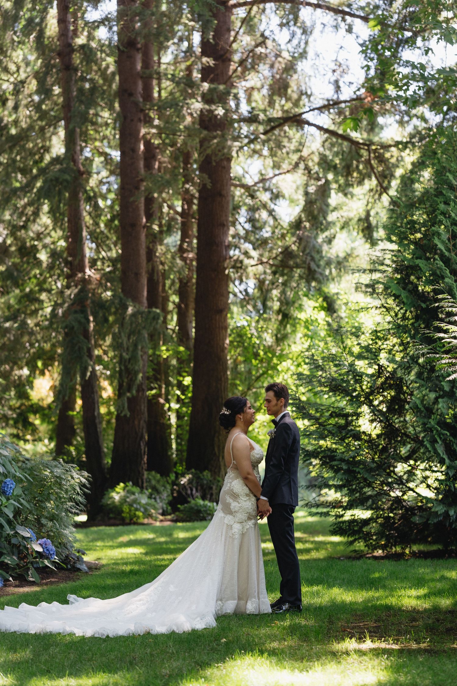 Bride and groom standing close together in a sunlit garden surrounded by tall evergreen trees.