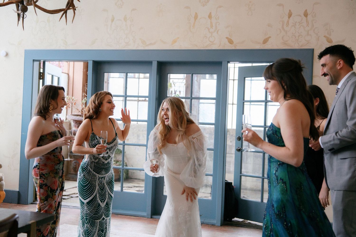 Bride laughing with bridesmaids in colorful dresses near blue French doors at wedding reception.