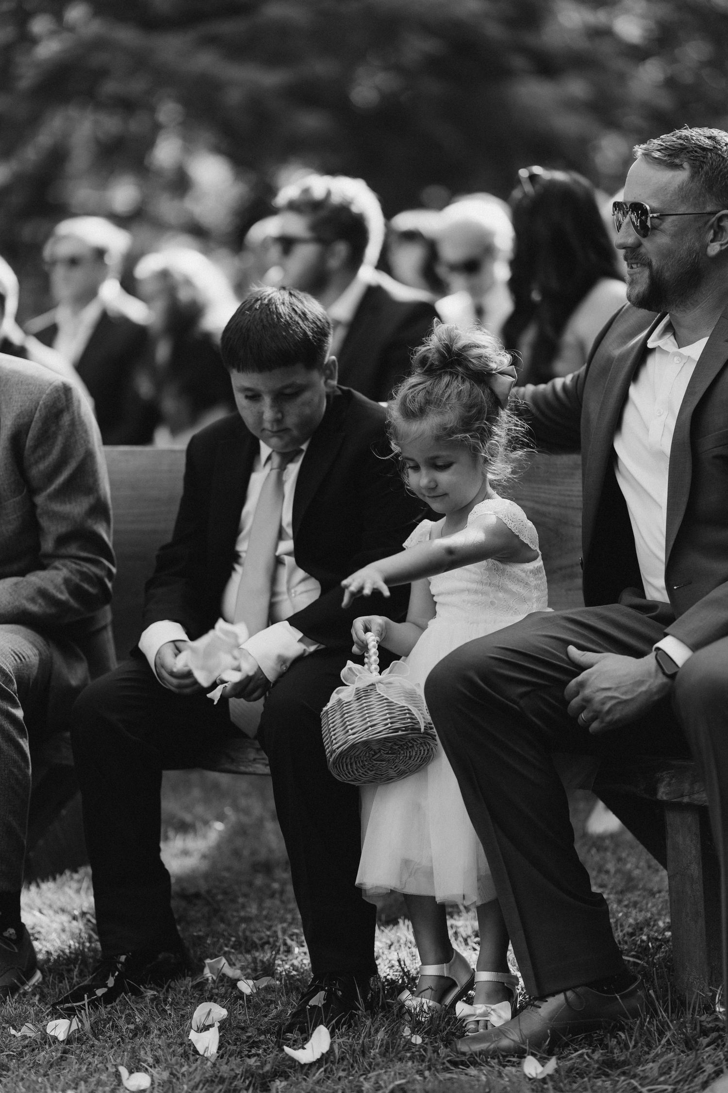 Black and white photo of flower girl and ring bearer sitting with father at outdoor wedding ceremony.