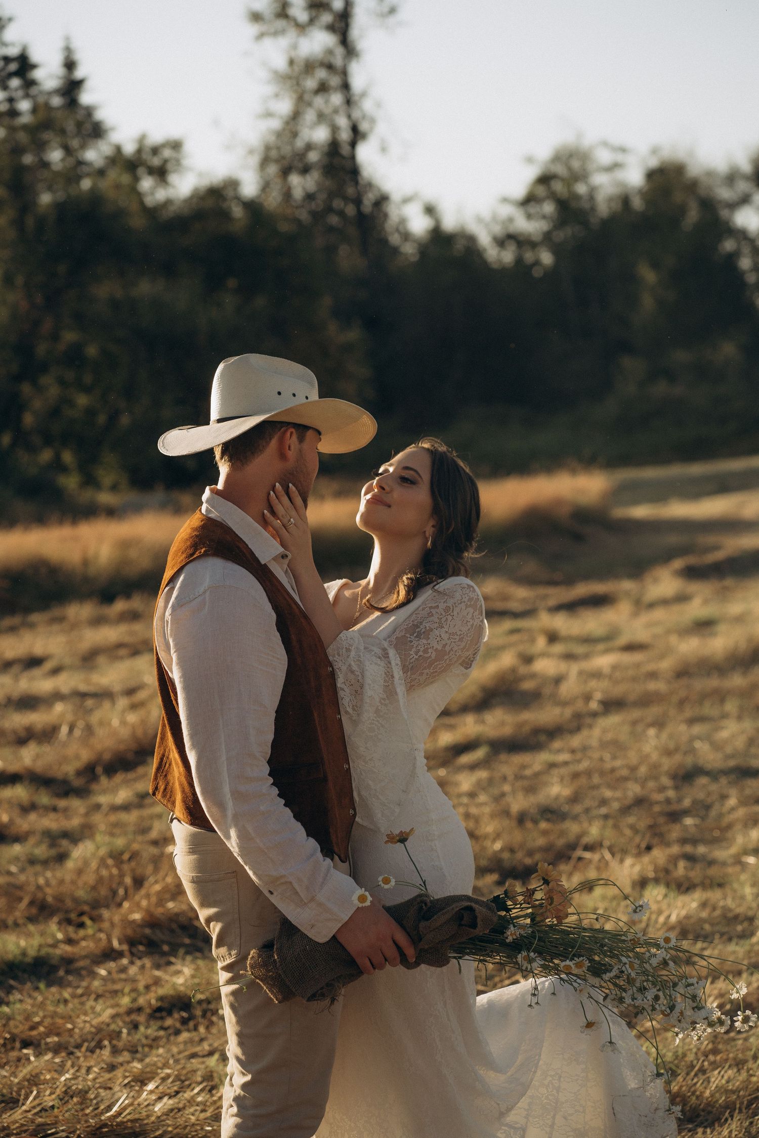 Bride gazing up at groom in cowboy hat while embracing in golden field at sunset.