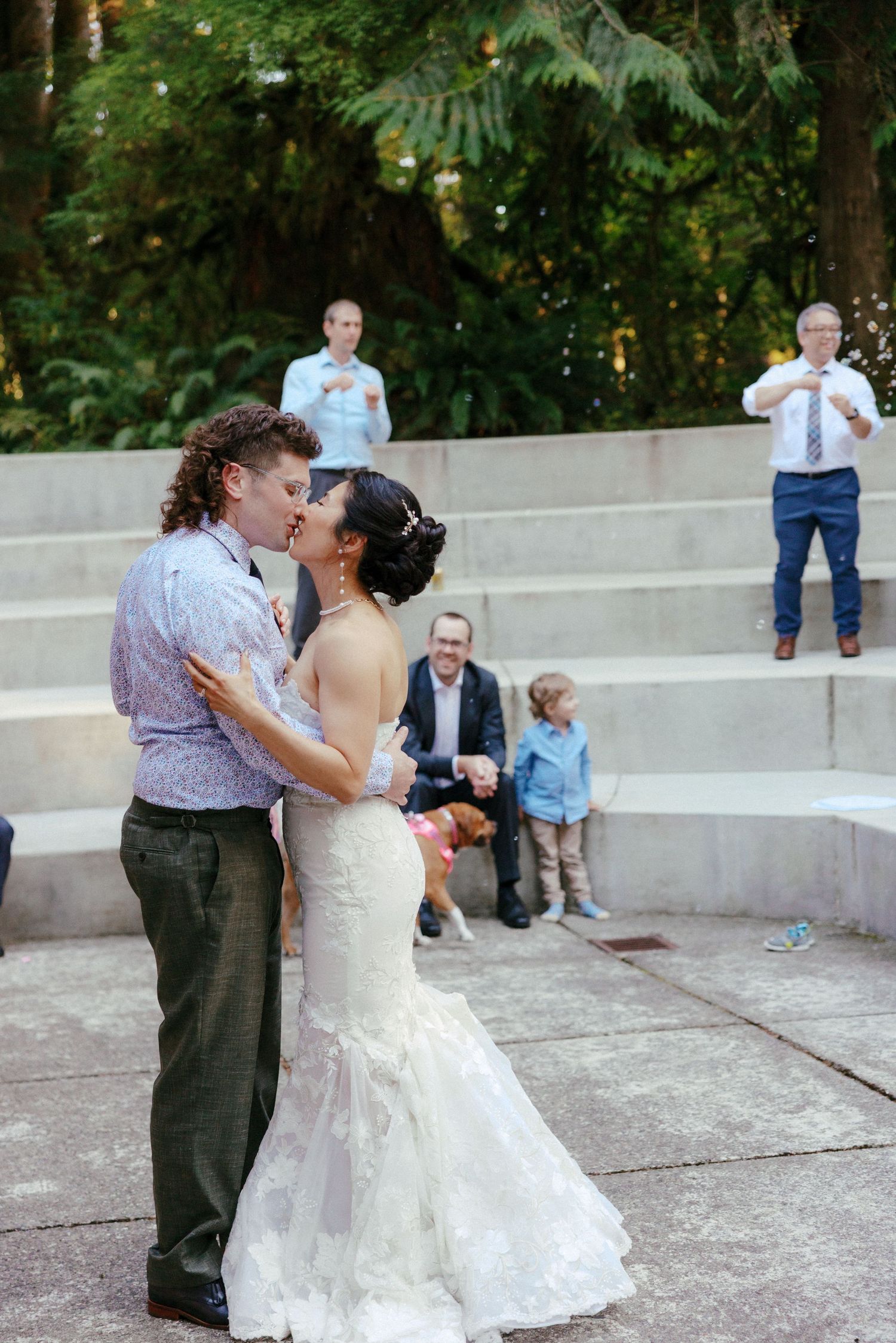 Bride and groom share first kiss at outdoor wedding while guests applaud on stone steps.