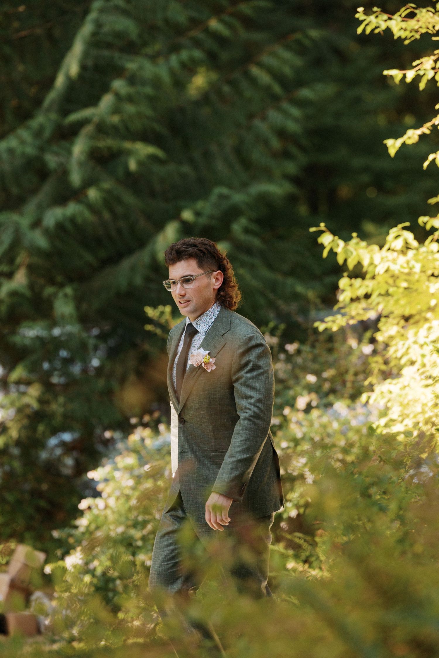 Dapper groom in olive gray suit with boutonniere walks through sunlit garden greenery at outdoor wedding.