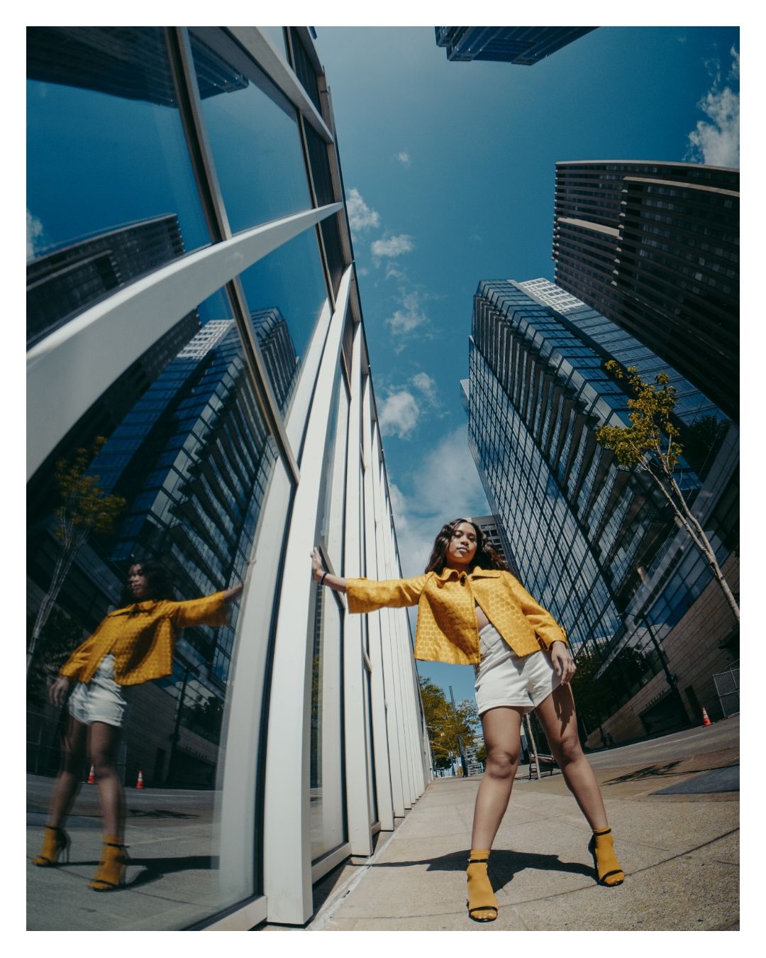 Woman in yellow jacket and white skirt poses confidently beside reflective glass skyscrapers using fisheye lens.