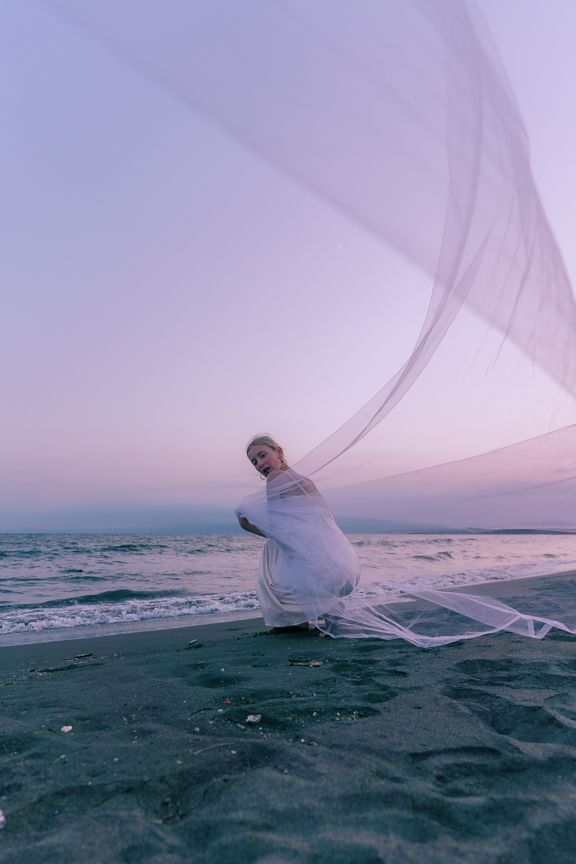 Woman in white dress crouches on sandy beach holding large flowing tulle fabric.