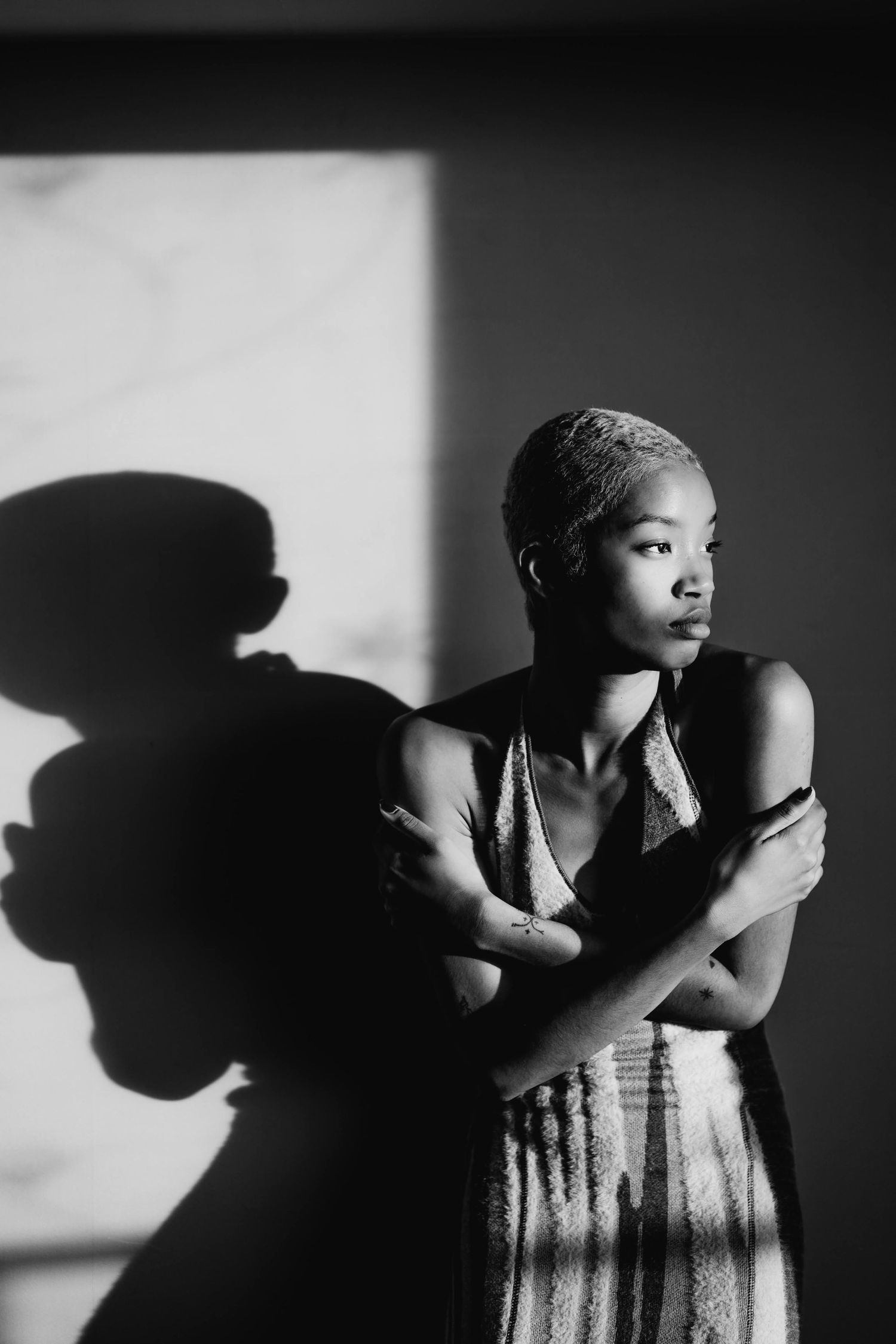 Black and white portrait of a woman with short hair crossing her arms, casting a dramatic shadow on the wall.