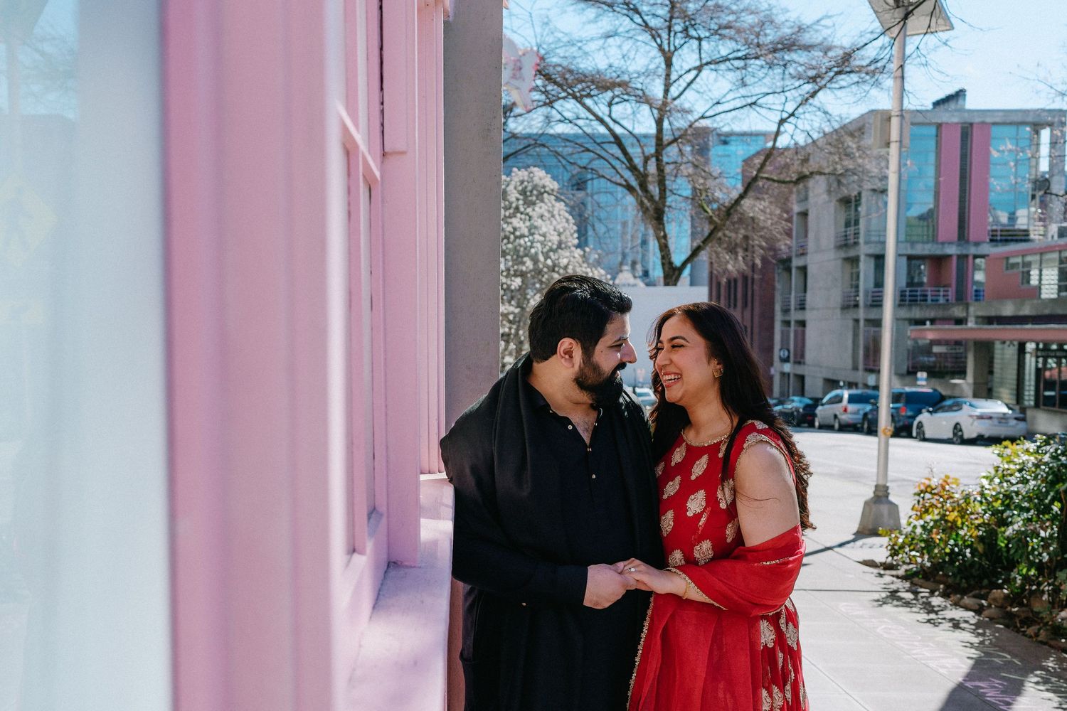 South Asian couple in traditional attire smiling at each other beside a pink building on a sunny day.