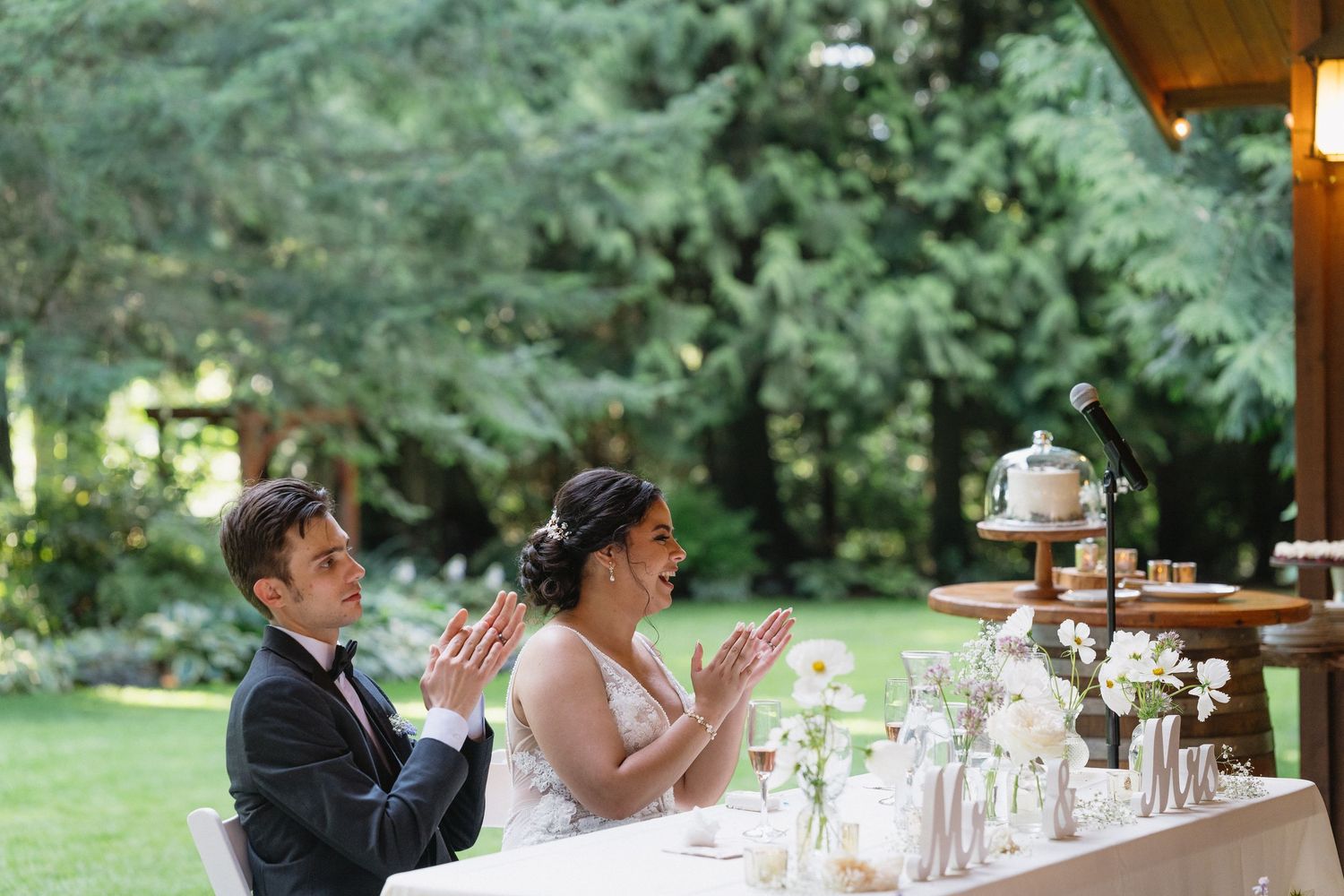 Bride and groom clapping at outdoor wedding reception sweetheart table decorated with white flowers.