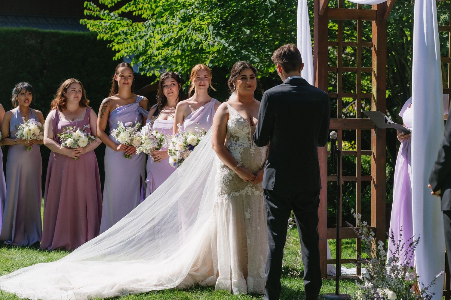 Bride in beaded gown faces groom at altar surrounded by bridesmaids in purple dresses with bouquets.
