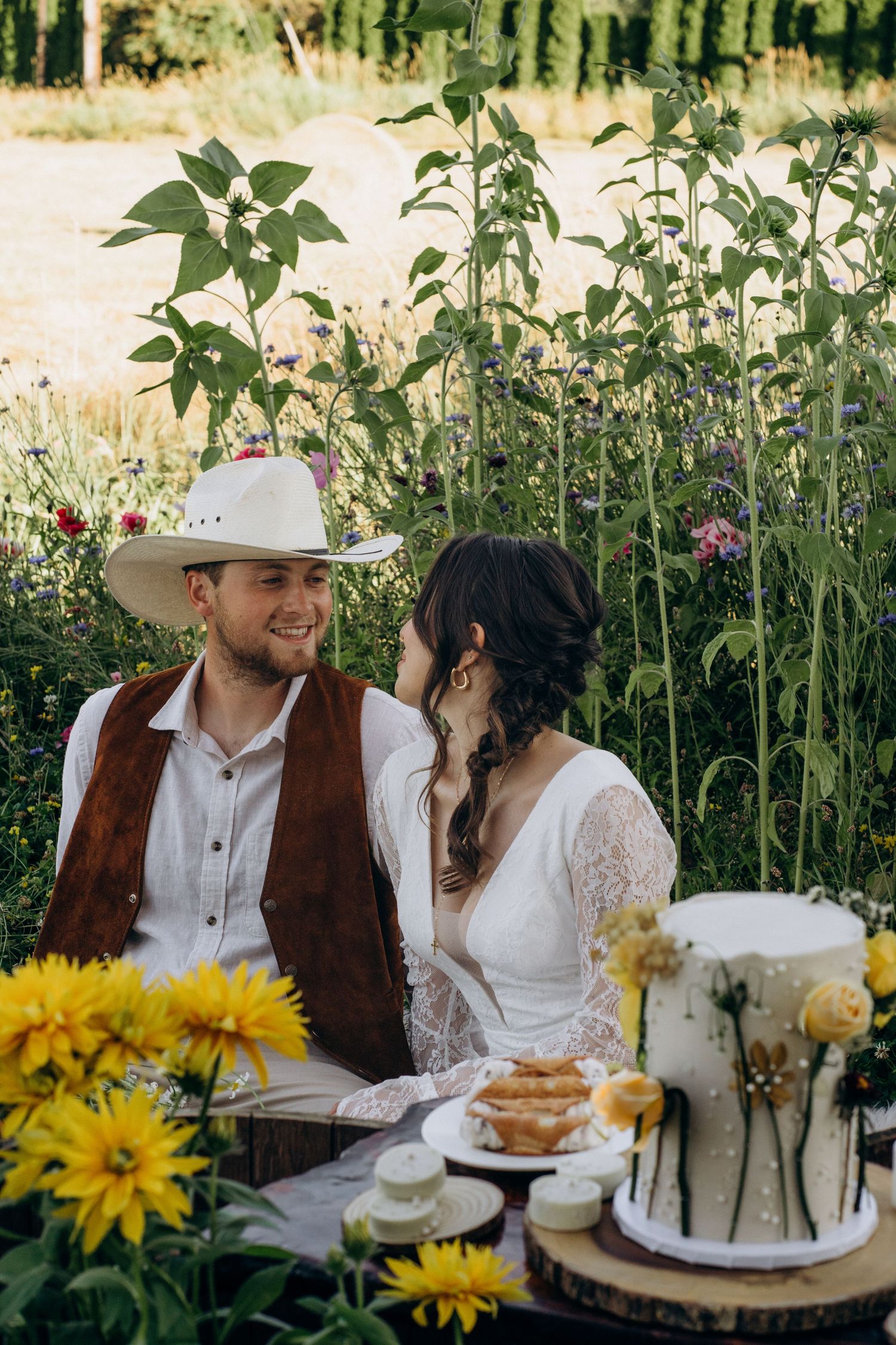 Smiling groom in cowboy hat and bride sit together near wedding cake in lush wildflower garden.