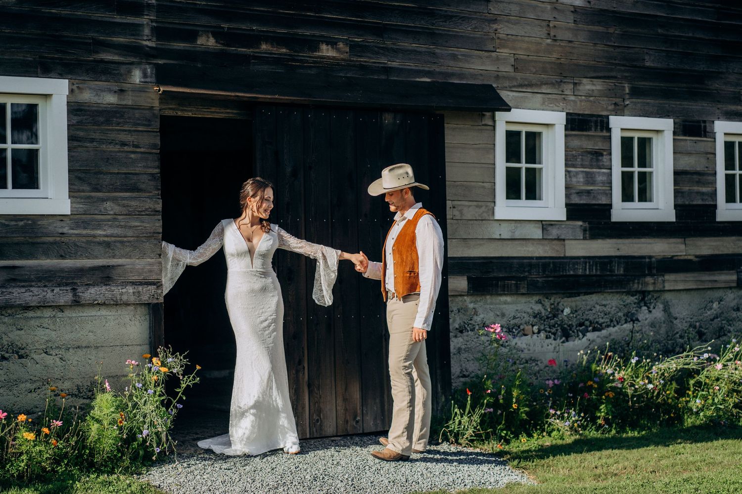 Bride and groom dancing together outside rustic barn surrounded by colorful wildflower garden.