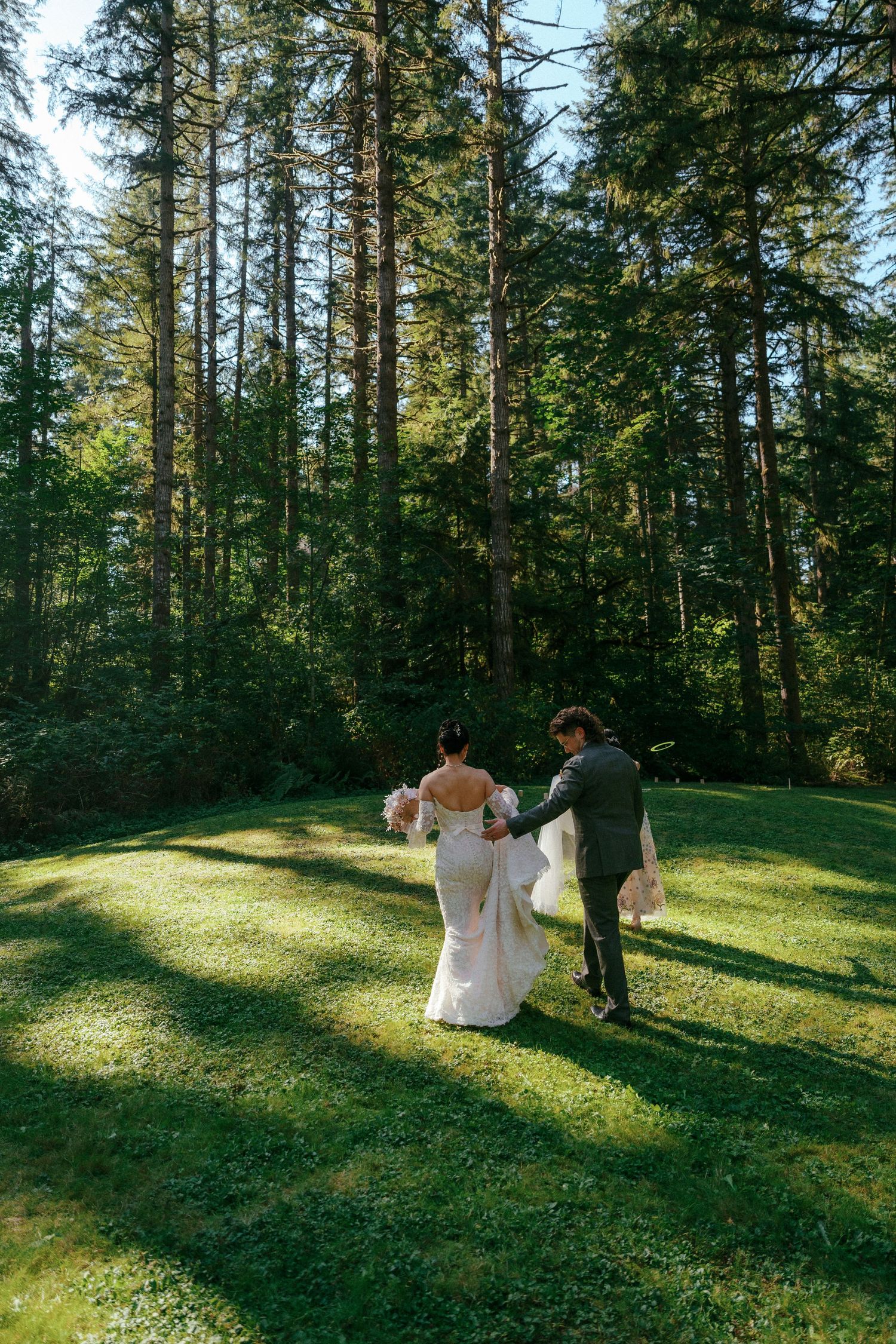 Bride and groom walk hand in hand across green lawn surrounded by tall trees.