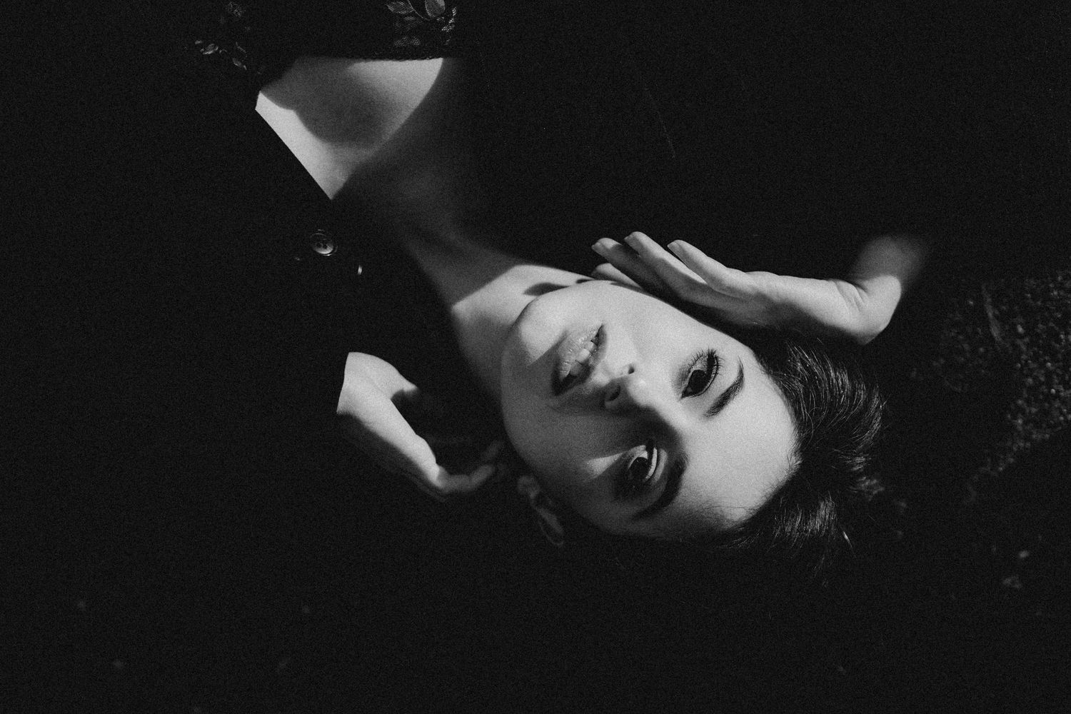 Black and white artistic portrait of woman lying back with hand raised against dark background.