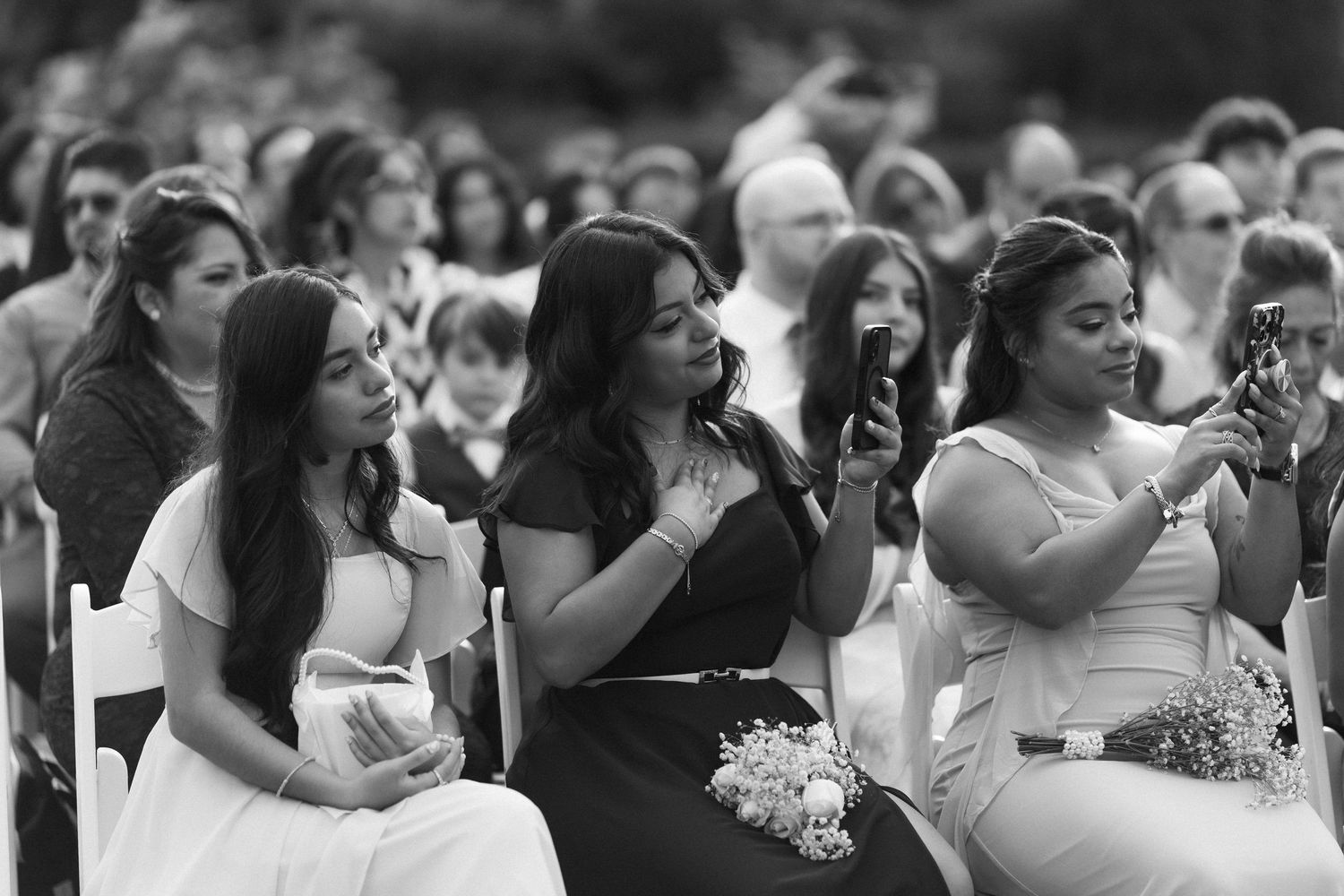 Wedding guests smile and capture moments on phones while seated at an outdoor wedding ceremony.
