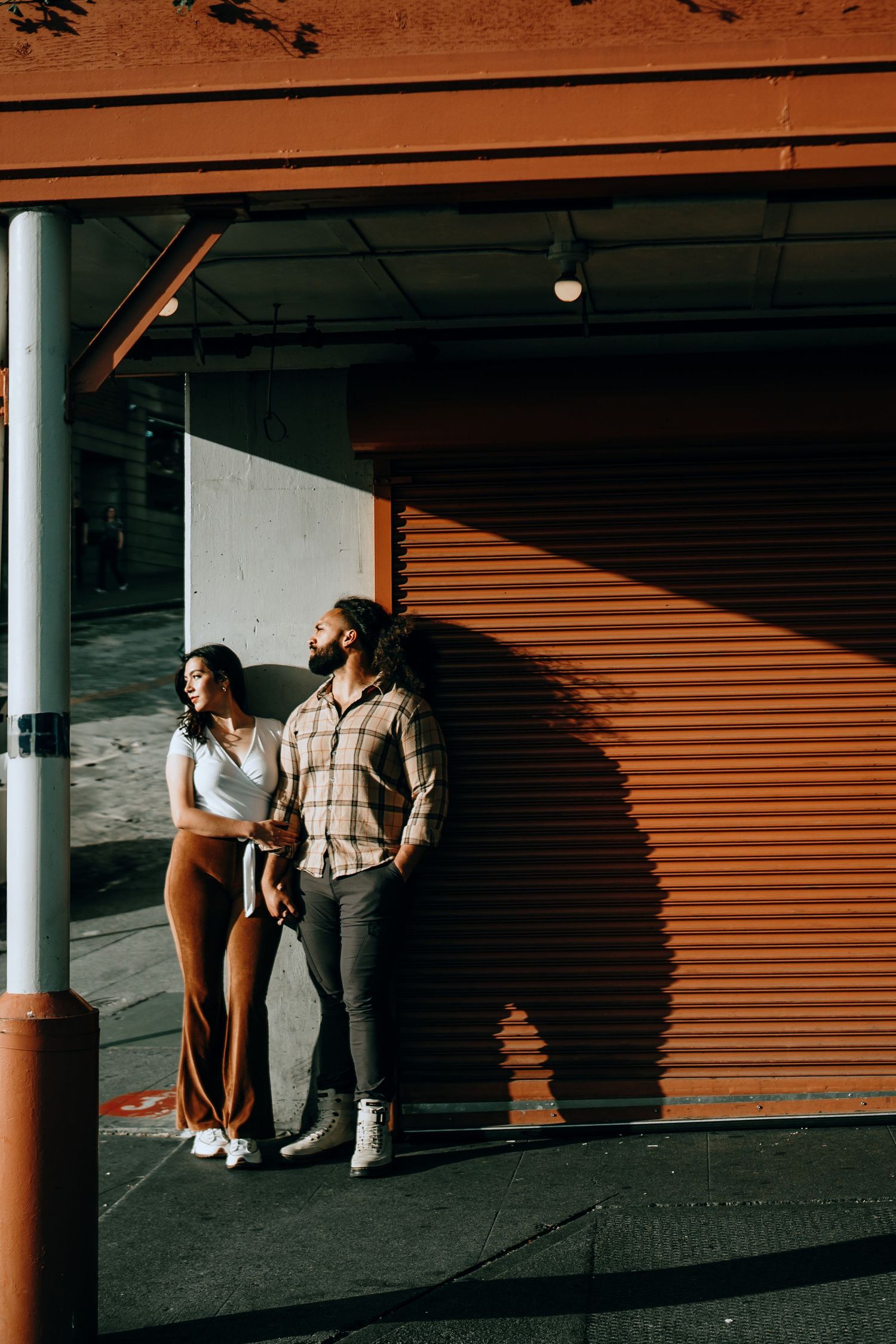 Stylish couple leaning against an orange corrugated wall in warm sunlight on a city street.